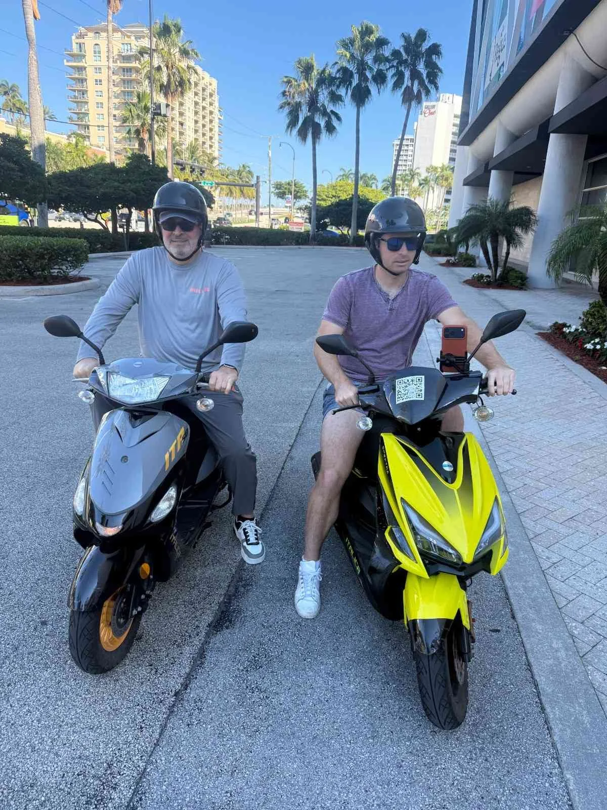 Two men wearing helmets and sunglasses sitting on electric scooters parked on a city street with palm trees, high-rise buildings, and a blue sky in the background.