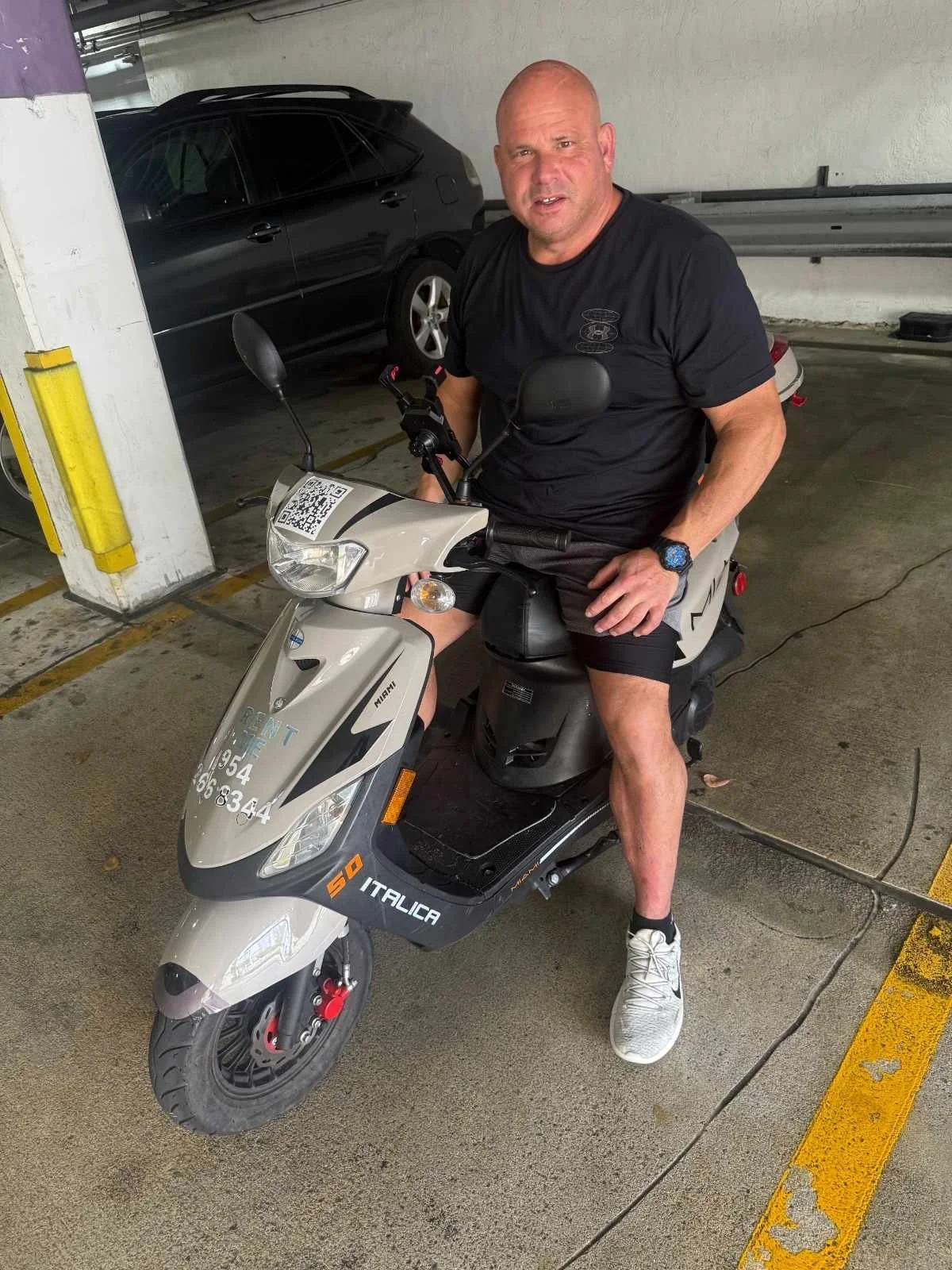 A man sitting on a small gray scooter in a parking garage, with a black car in the background. The man is wearing a black T-shirt, black shorts, white sneakers, and a blue watch.