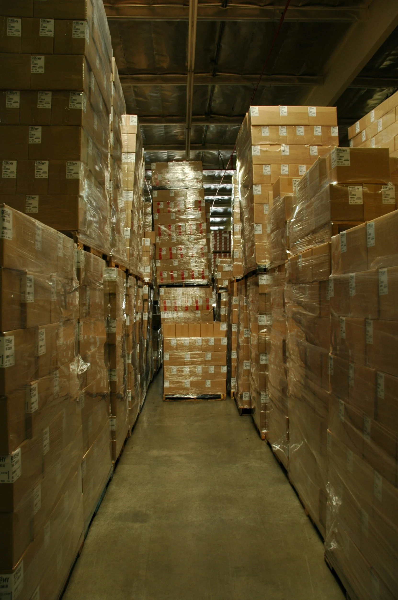 A warehouse aisle filled with stacked cardboard boxes wrapped in clear plastic, stored on pallets along the sides and in the middle.