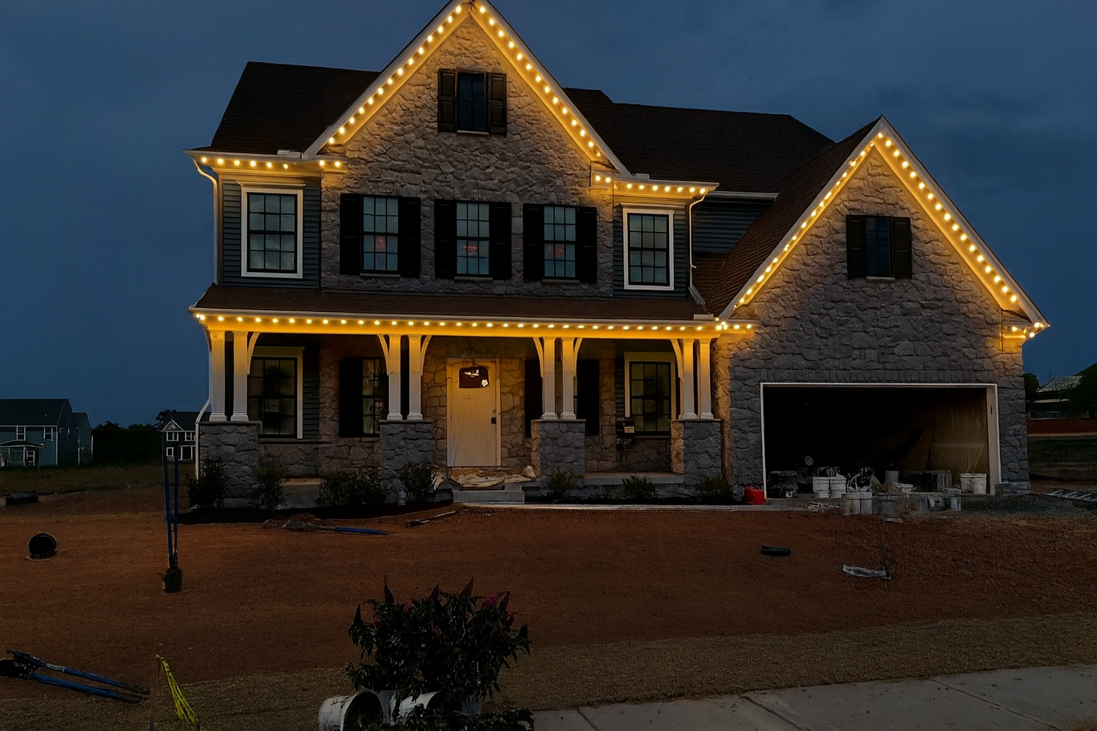 A two-story house at night with exterior lights, a garage door, a front porch with chairs, and a wreath on the door, surrounded by a lawn.