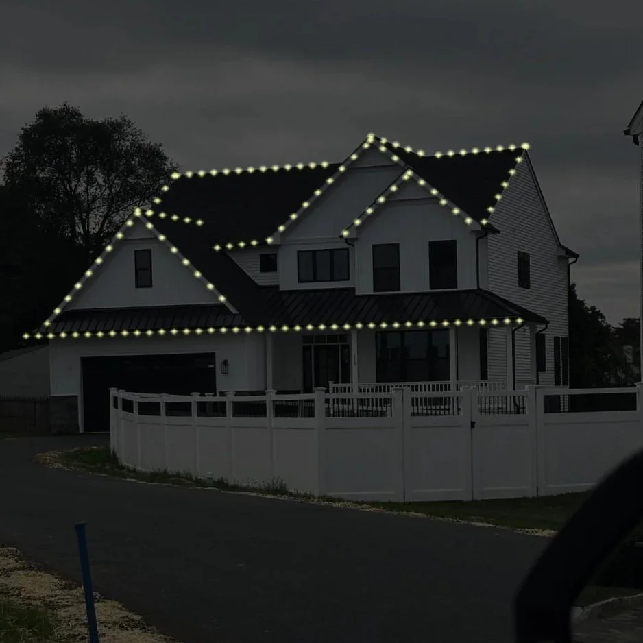 A two-story house decorated with white Christmas lights outlining the roof and windows at night. The house has black shutters, a front door with a wreath, and a garage door. The yard has bushes and trees also decorated with white lights, and a dark sky serves as the background.