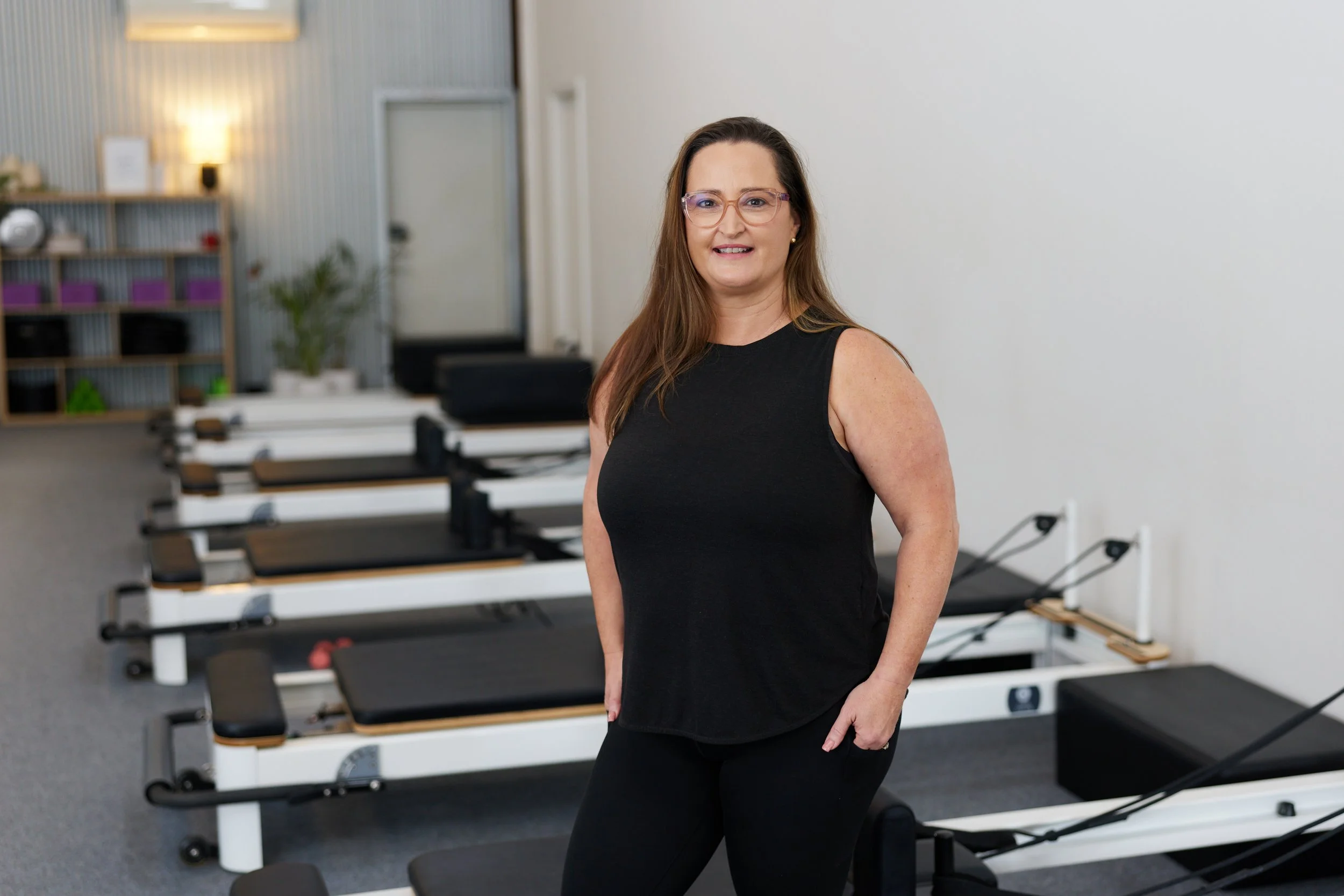 A woman with long brown hair, wearing glasses and a black sleeveless top, stands in a fitness or physical therapy studio with Pilates reformer machines and workout equipment in the background.