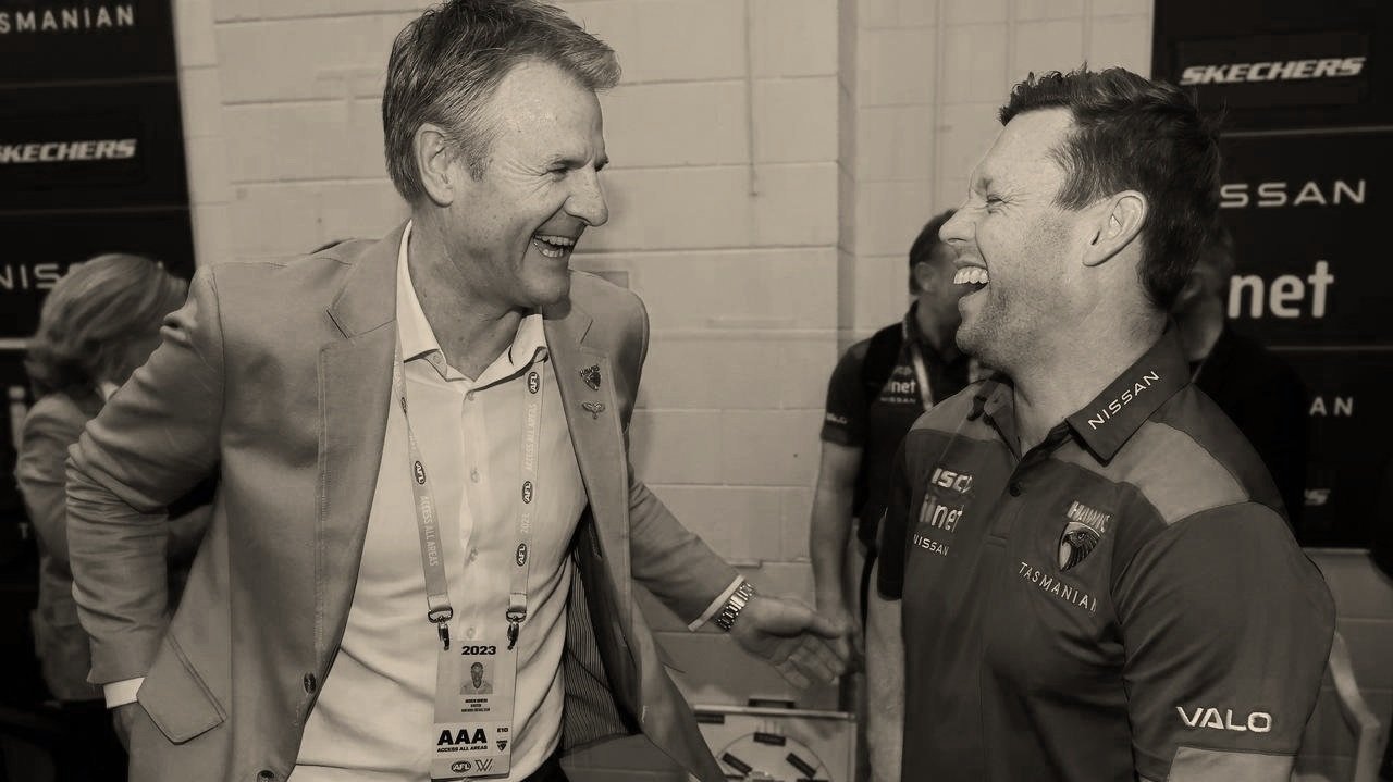 Two men laughing and conversing, one in a suit with event badge and the other in a branded sports shirt, with sponsors' logos in the background.