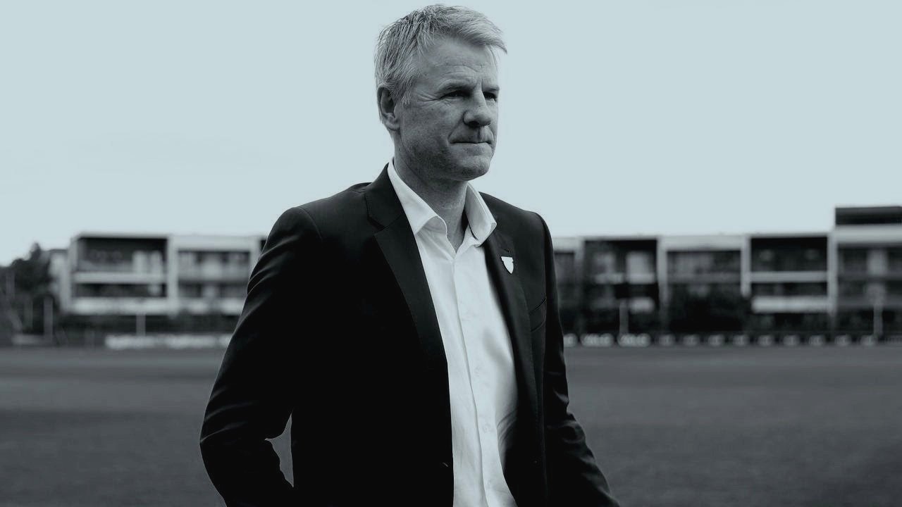 A grayscale image of a man in a suit standing outdoors with buildings in the background.