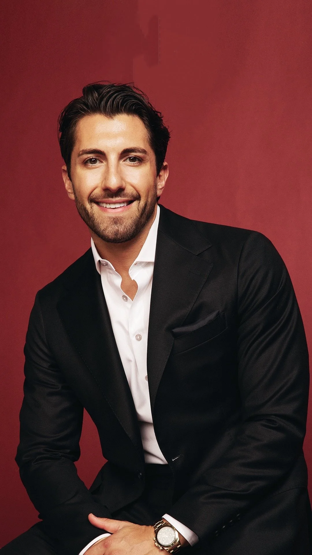 Jason Tartick in a black suit with a white shirt sitting against a red background
