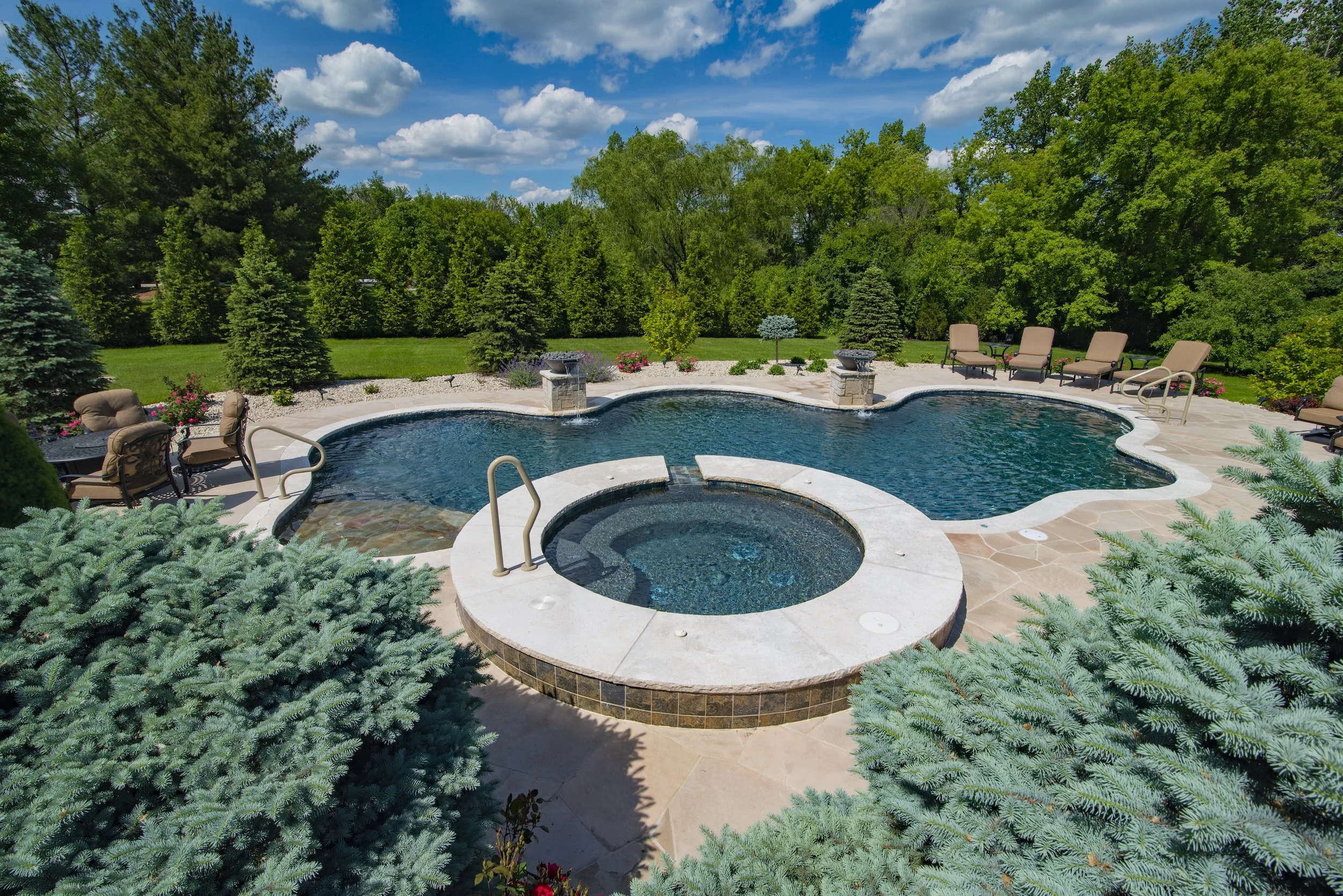 Outdoor swimming pool with circular hot tub and patio chairs, surrounded by trees and greenery.
