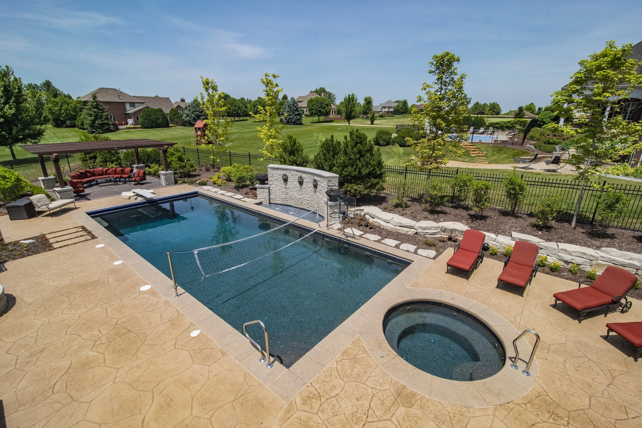Backyard with a swimming pool, spa, and lounge chairs. A net spans the pool for volleyball, adjacent to a stone wall with water features. There's a pergola with seating next to landscaped greenery and a large lawn. Suburban houses and trees are visib
