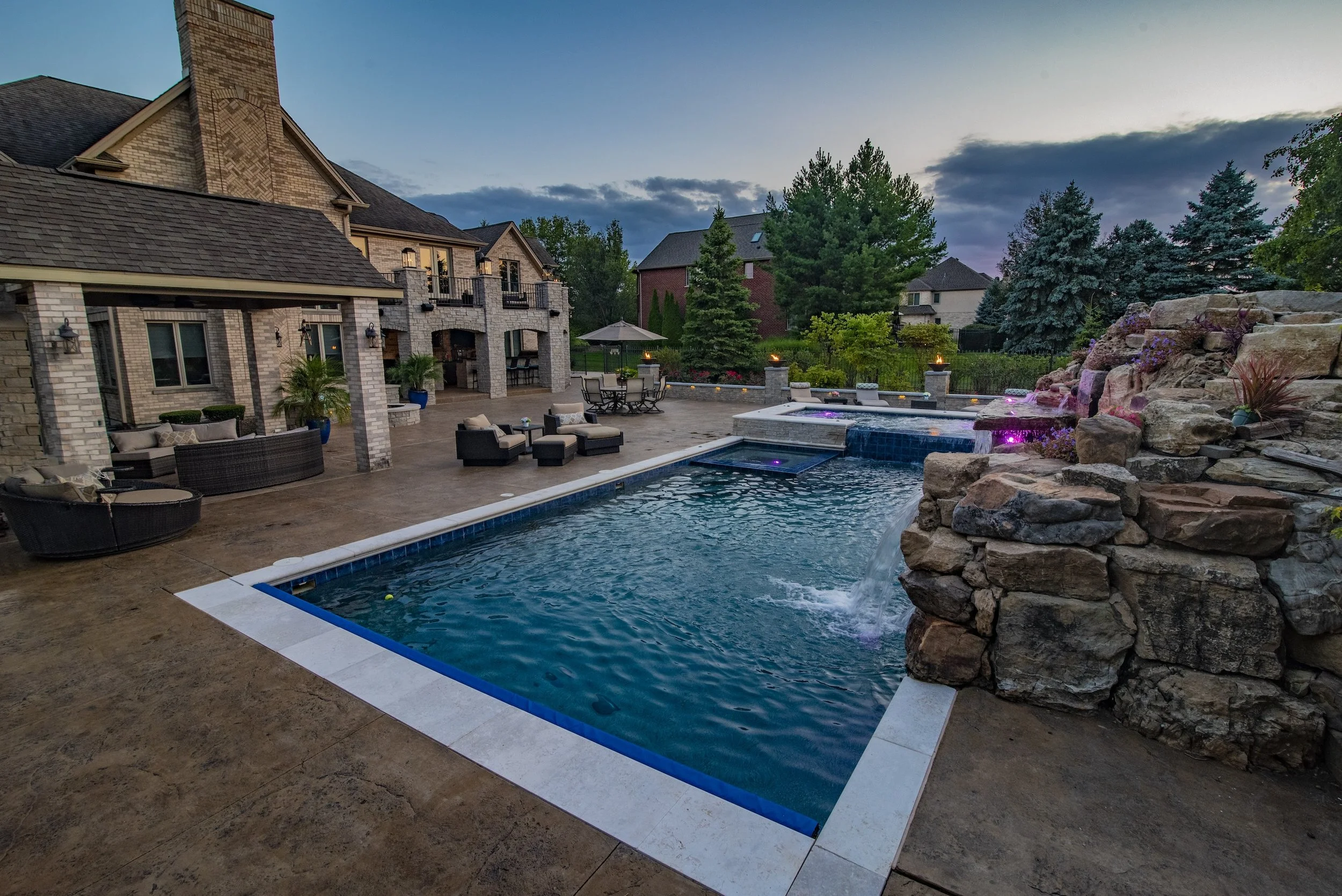 Luxury backyard with a pool, waterfall, and patio seating adjacent to a large brick house, surrounded by landscaped trees and greenery.