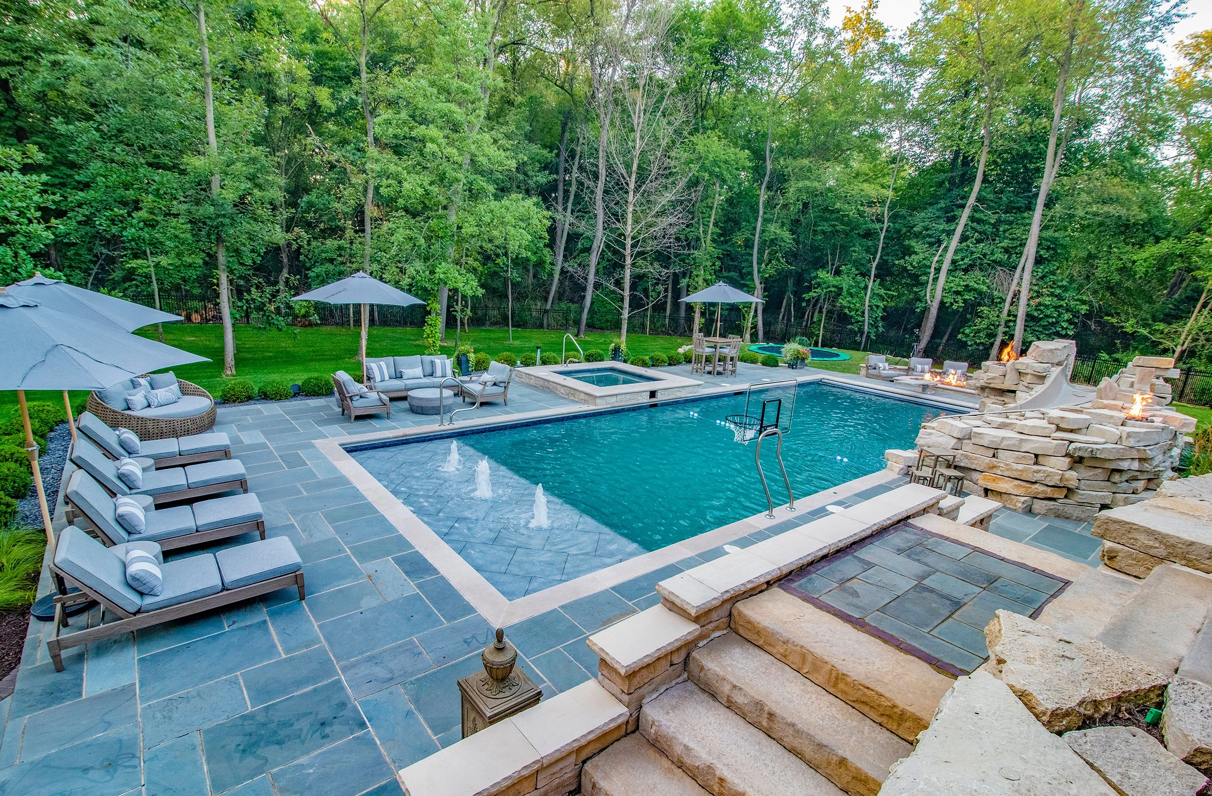 Backyard with a swimming pool, stone patio, lounge chairs, umbrellas, and a slide. Fire features and seating areas are around the pool, with a lush forest backdrop.