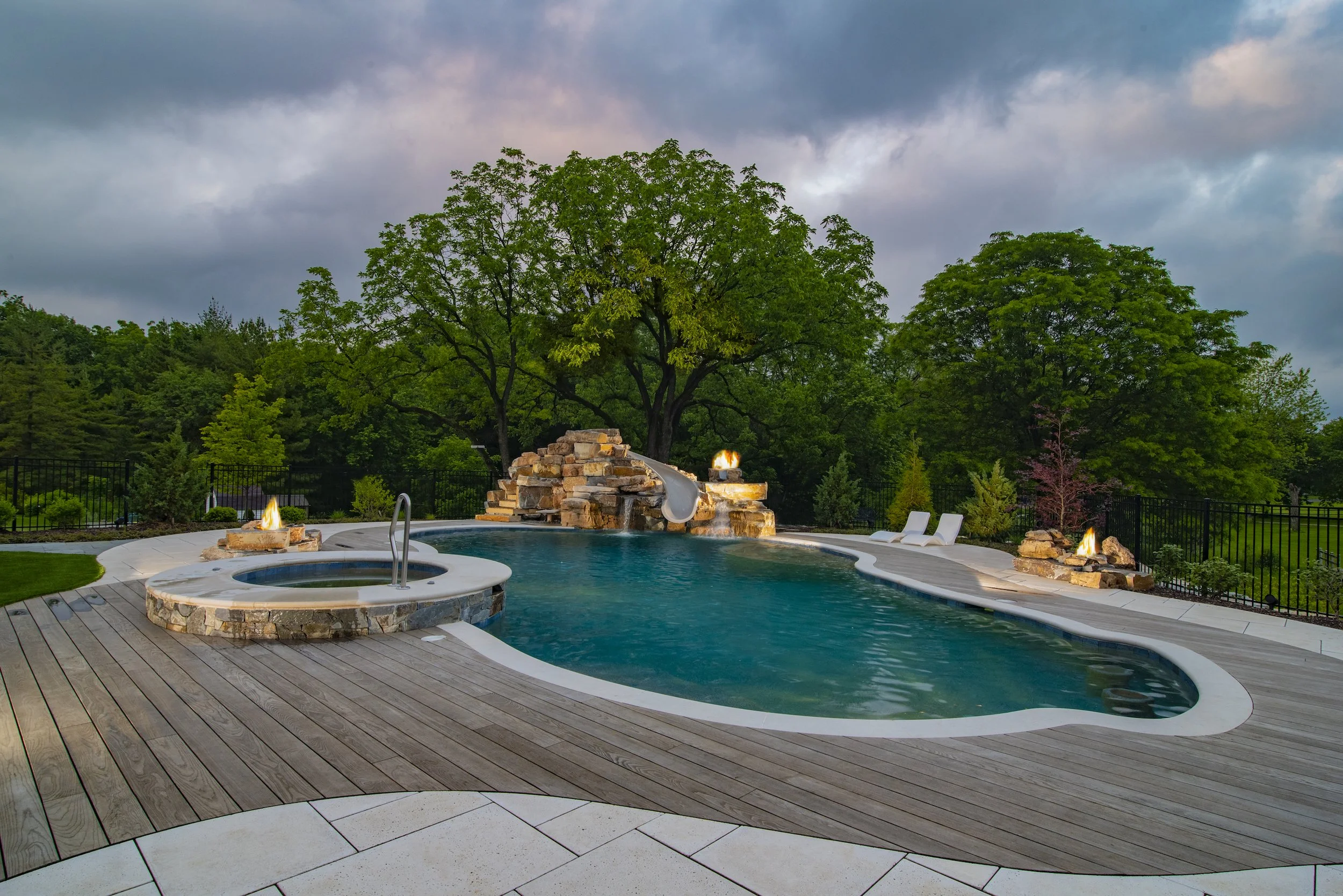 Outdoor swimming pool with stone waterfall, wooden deck, fire pits, and surrounding trees under a cloudy sky.