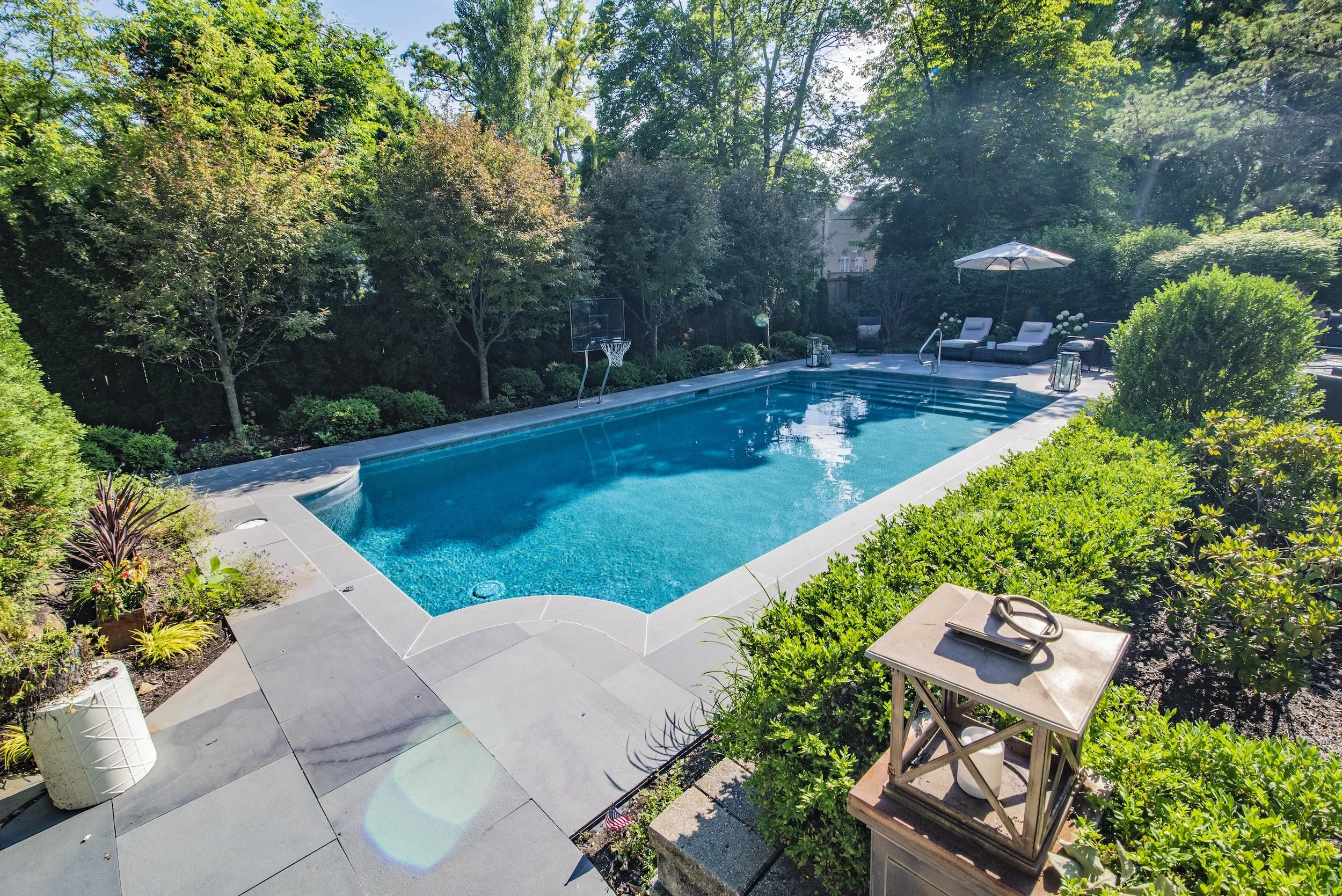 A backyard swimming pool surrounded by lush greenery and trees, with a stone patio. There is a poolside basketball hoop and lounge chairs with an umbrella. A decorative lantern sits on a pillar in the foreground.