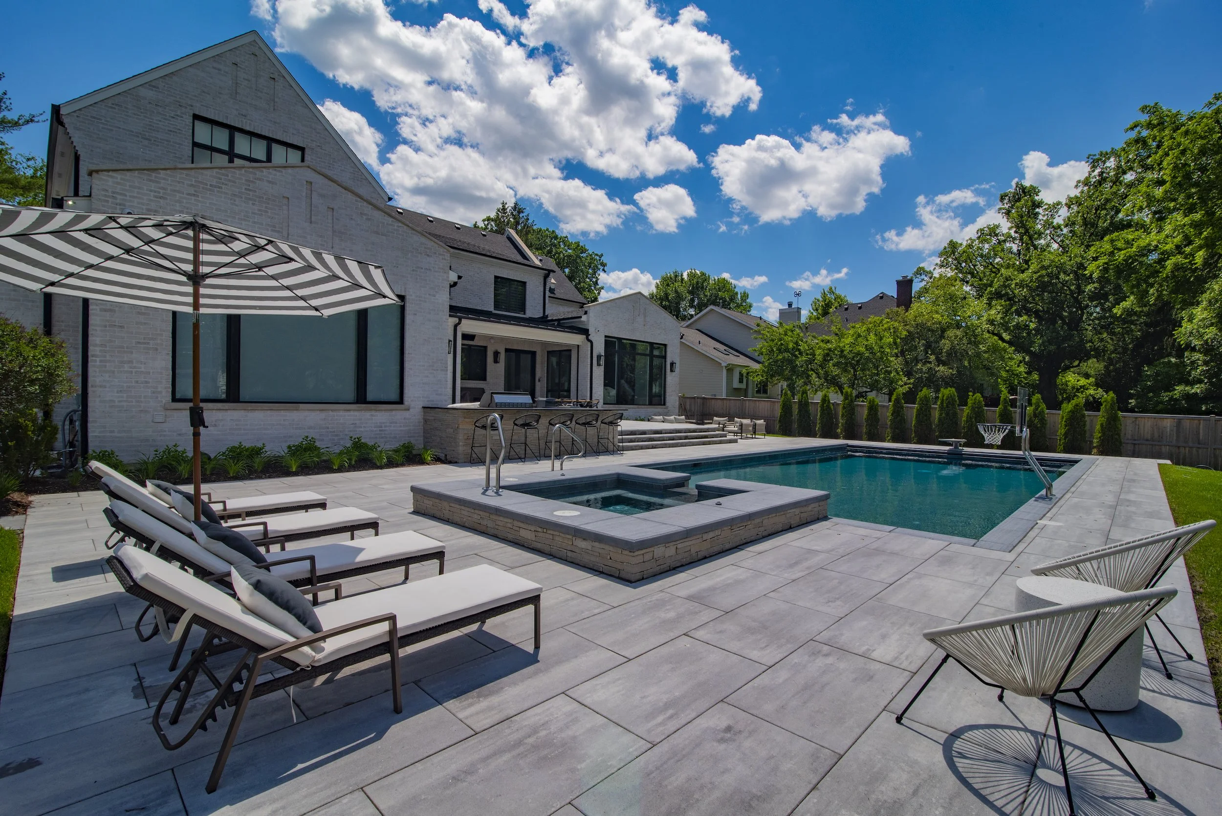 Modern backyard with a rectangular swimming pool, hot tub, lounge chairs under an umbrella, and a white brick house in the background.