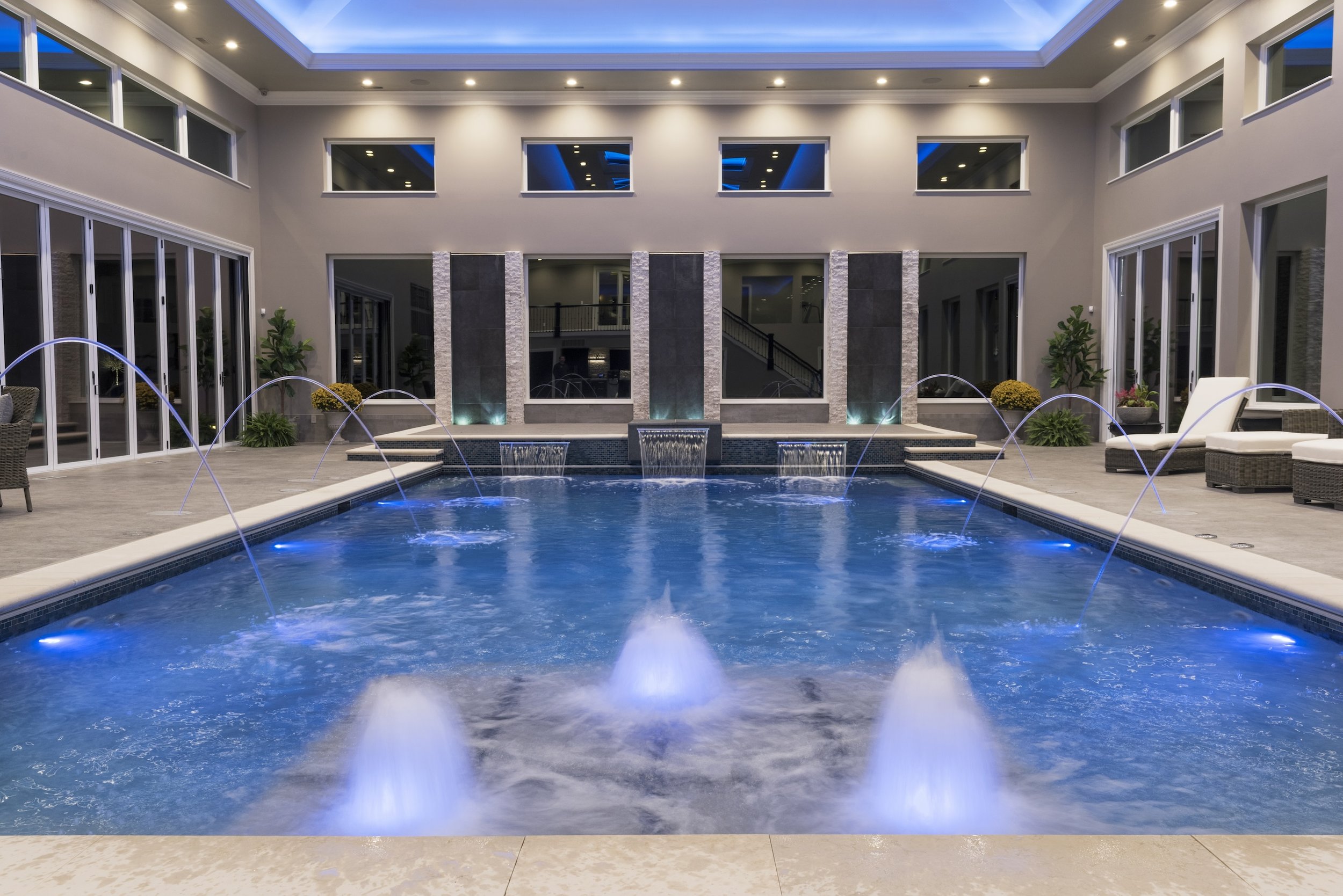 Indoor swimming pool with water fountains and modern lighting, surrounded by lounge chairs and potted plants.