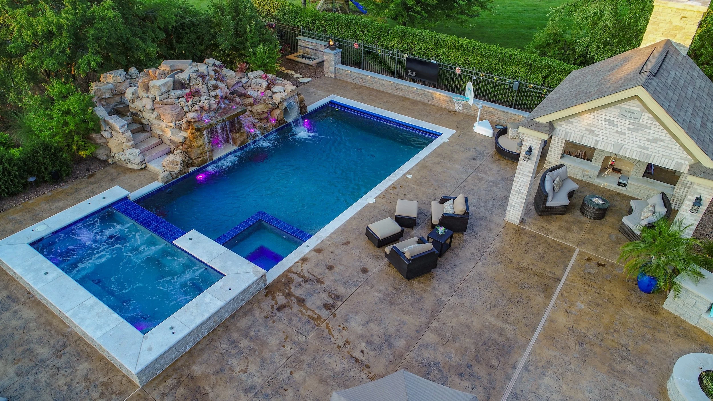 Aerial view of a backyard with a swimming pool, hot tub, waterfall feature, poolside seating, and a covered patio area with outdoor furniture.