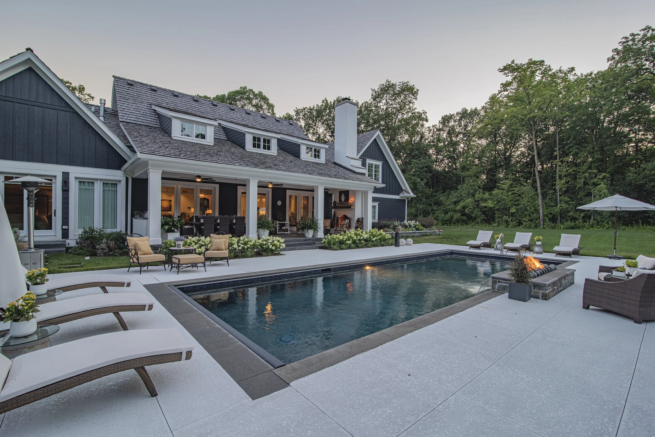 Backyard with swimming pool, luxury house, patio seating, and fire pit surrounded by greenery and trees.