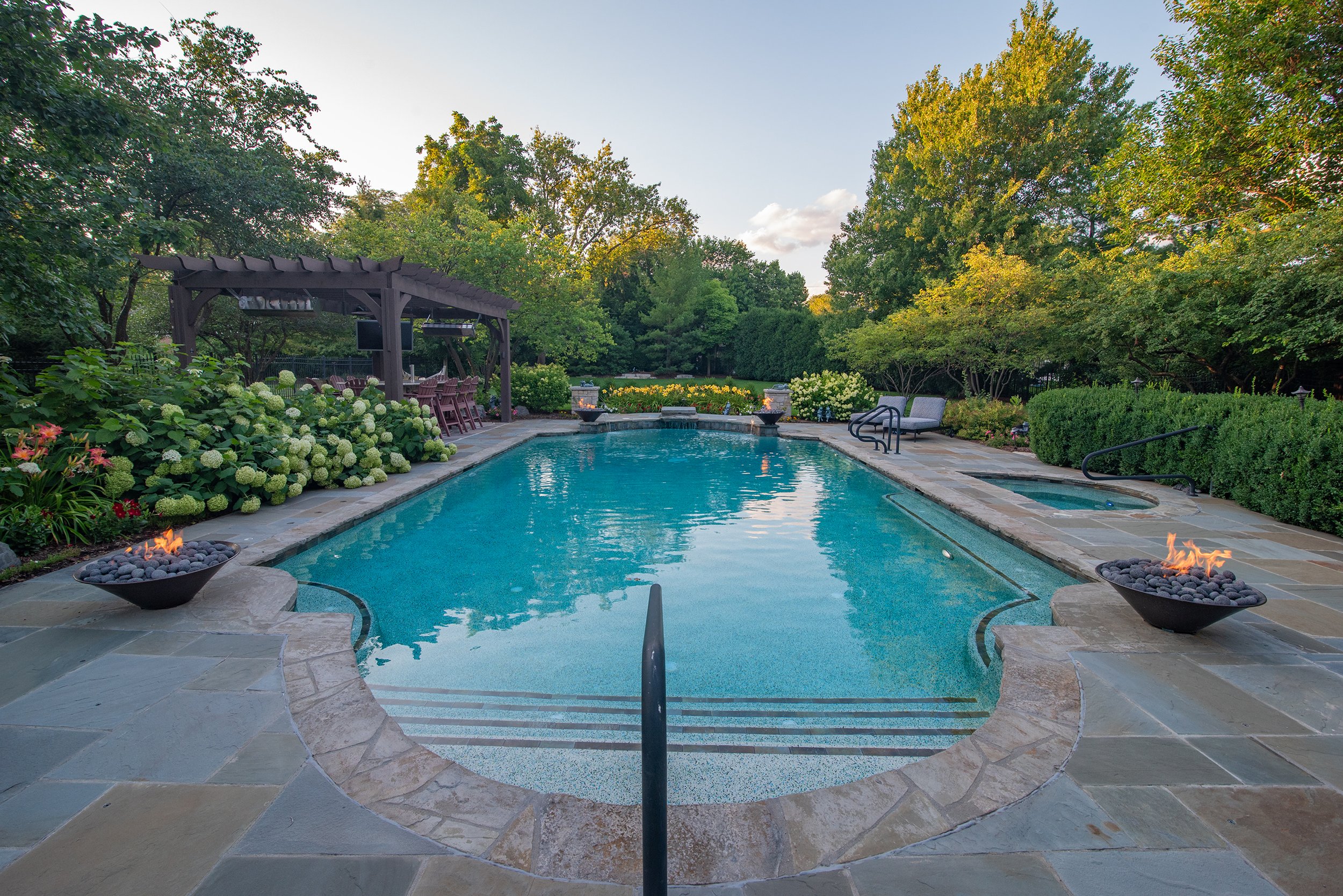 Backyard with a swimming pool, stone patio, fire bowls, pergola, outdoor dining area, lounge chairs, and lush landscaping with trees and flowers.