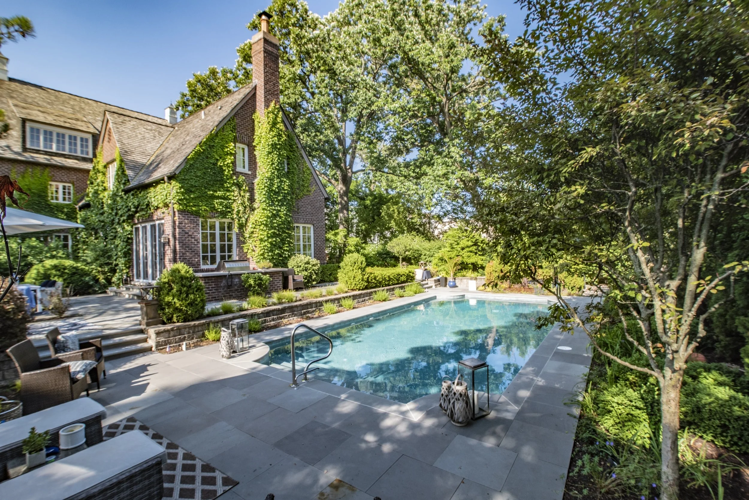 Brick house with ivy, surrounded by lush greenery, features a rectangular swimming pool on a sunny day.