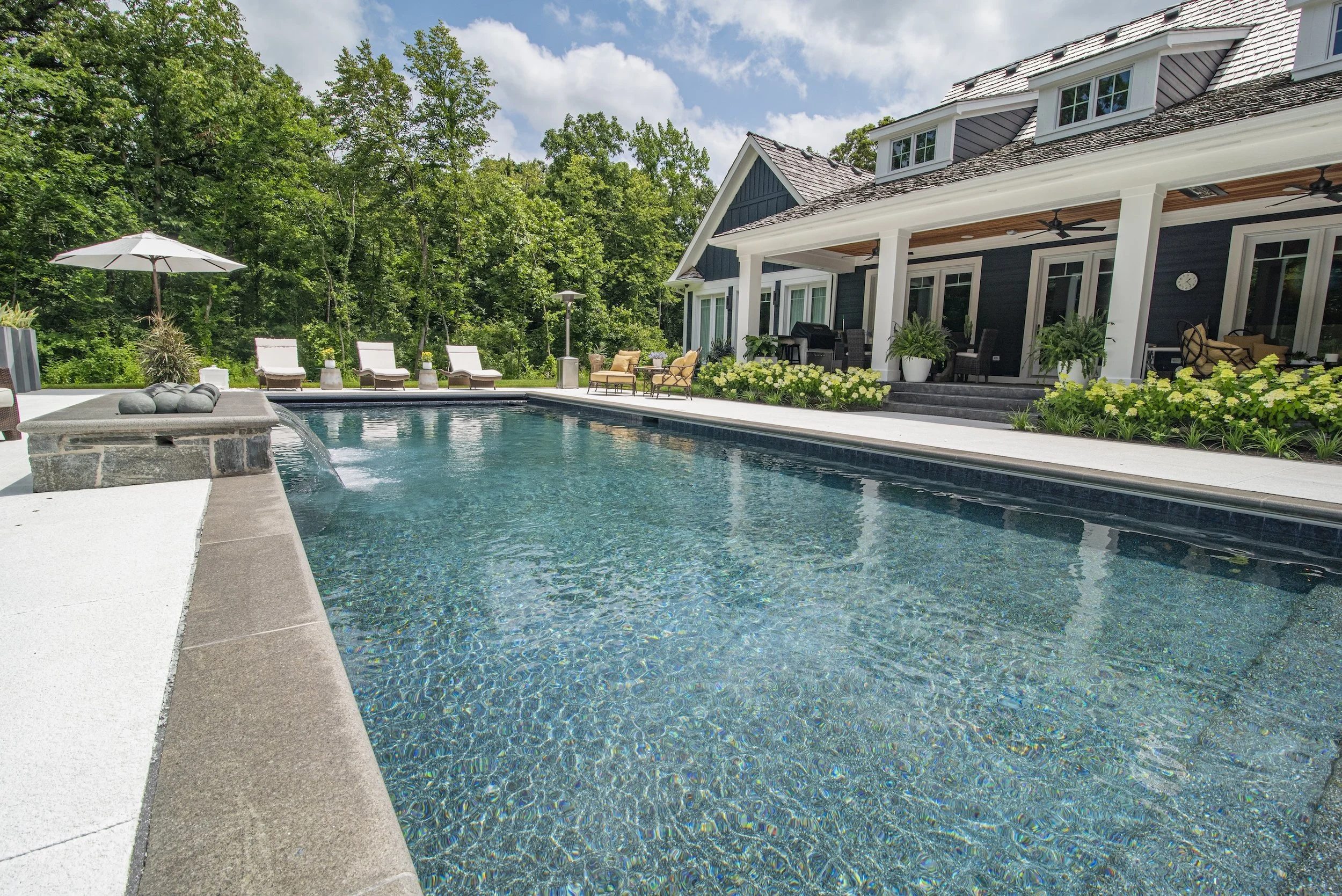 Luxury outdoor pool with lounge chairs, patio furniture, and lush greenery, adjacent to a modern house.