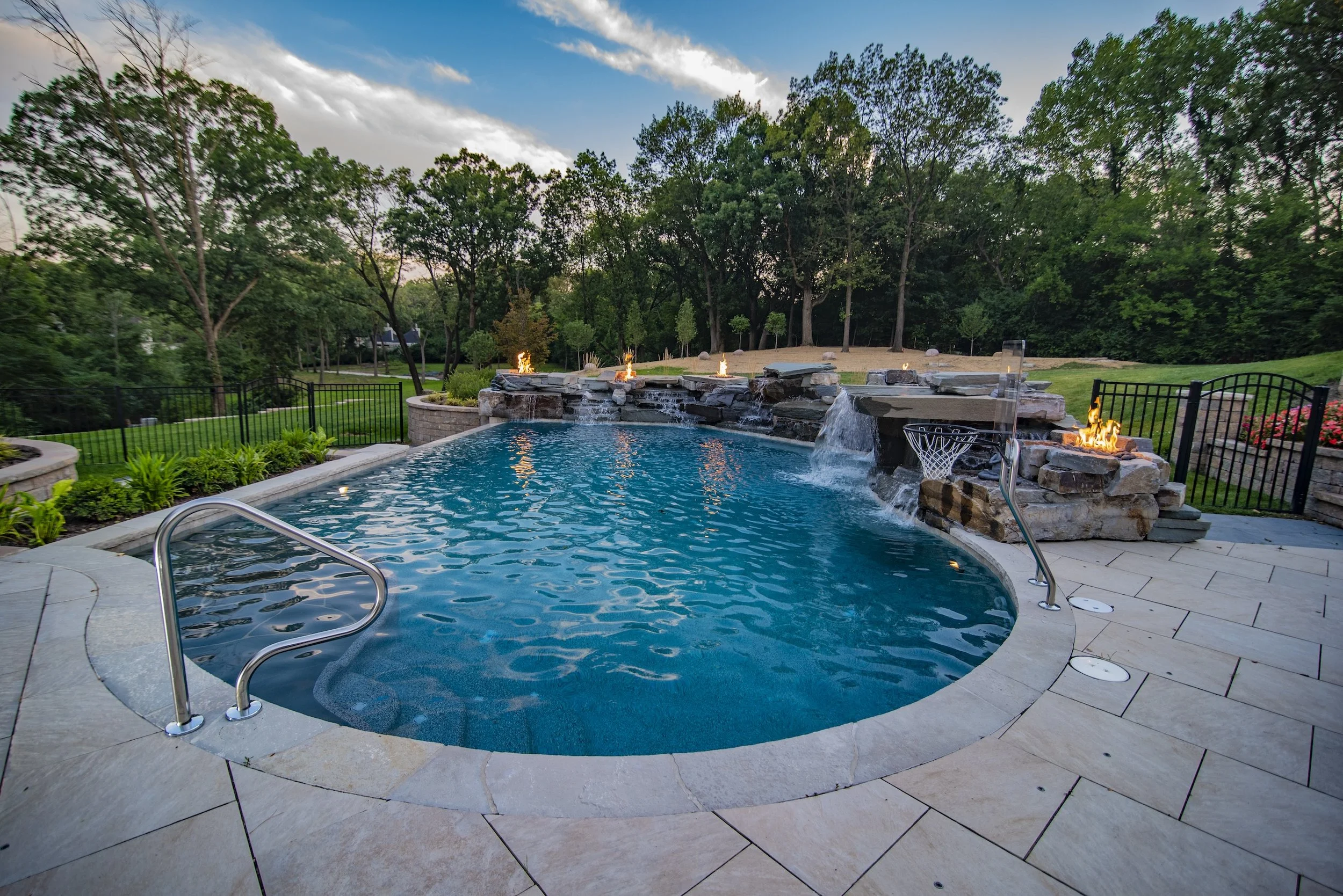 Luxury backyard pool with waterfalls, fire features, and a basketball hoop, surrounded by trees and a metal fence.