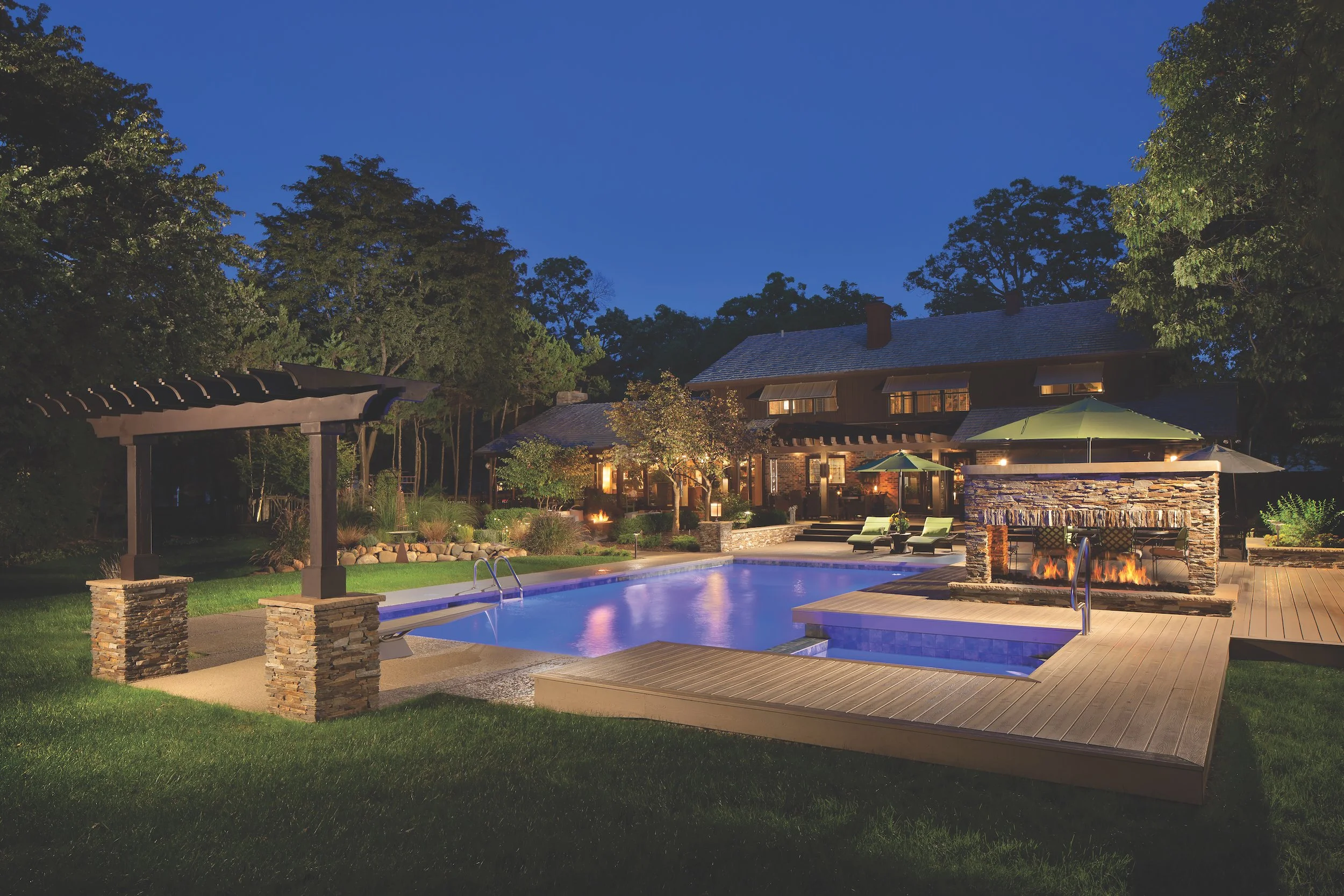 Luxurious backyard with illuminated swimming pool, pergola, fire feature, patio seating, and large house in the background, surrounded by trees in the evening.
