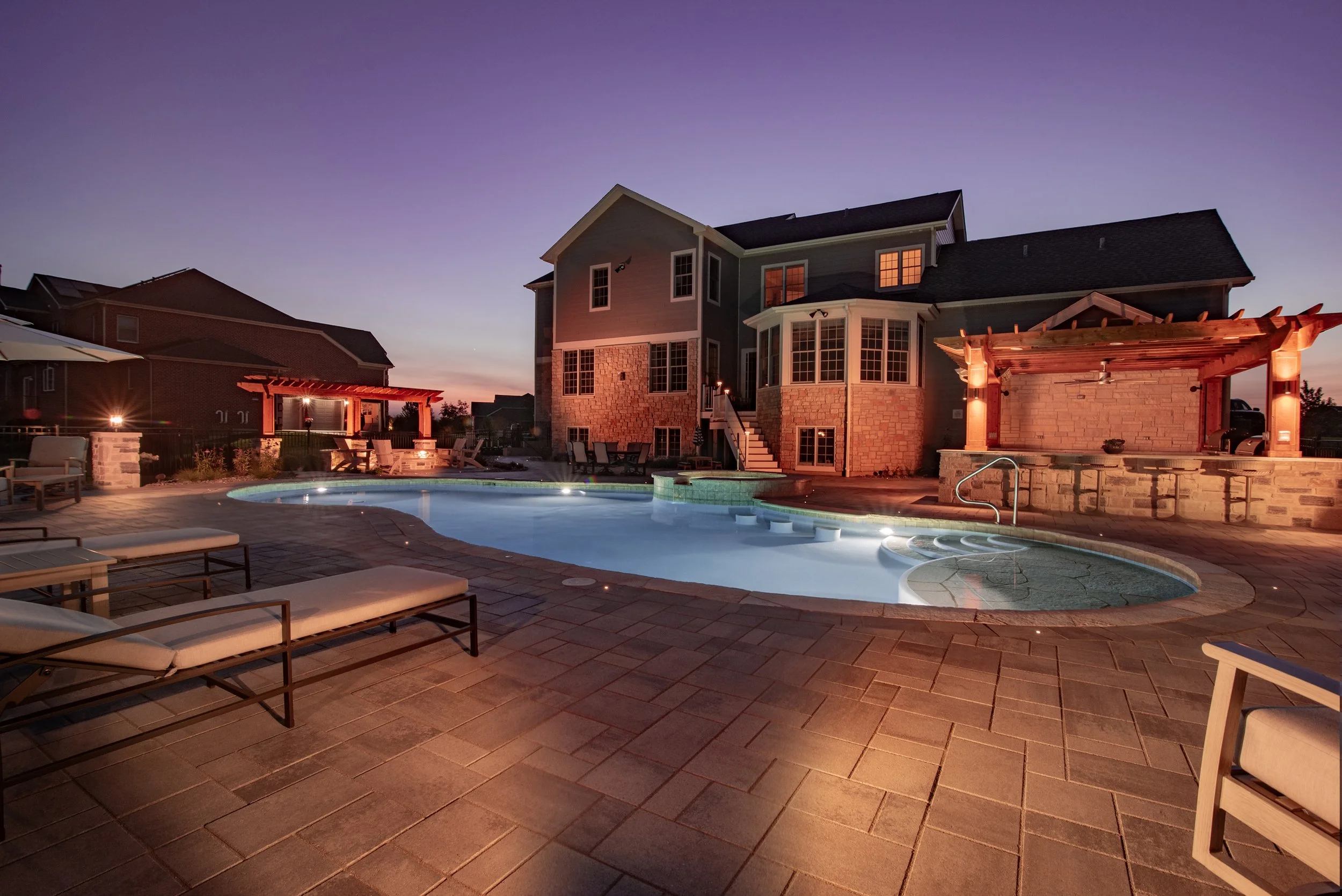 Modern backyard with illuminated swimming pool, deck chairs, and a pergola near a large house at dusk.
