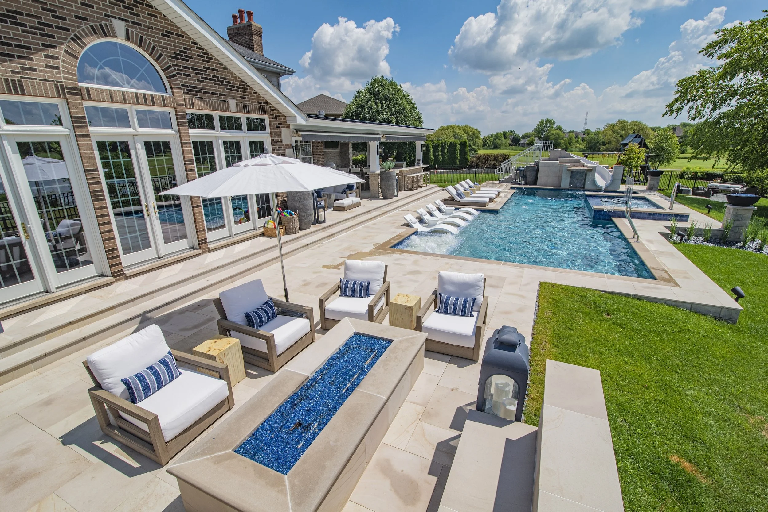 Luxury backyard with a swimming pool, lounge chairs, patio furniture, and a fire pit, adjacent to a brick house with large windows, under a clear blue sky.