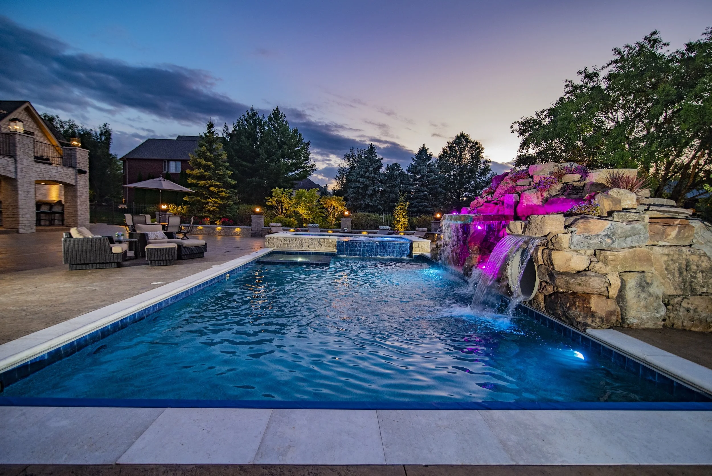 Luxurious backyard swimming pool with rock waterfall and colored lights, surrounded by patio furniture and trees at dusk.