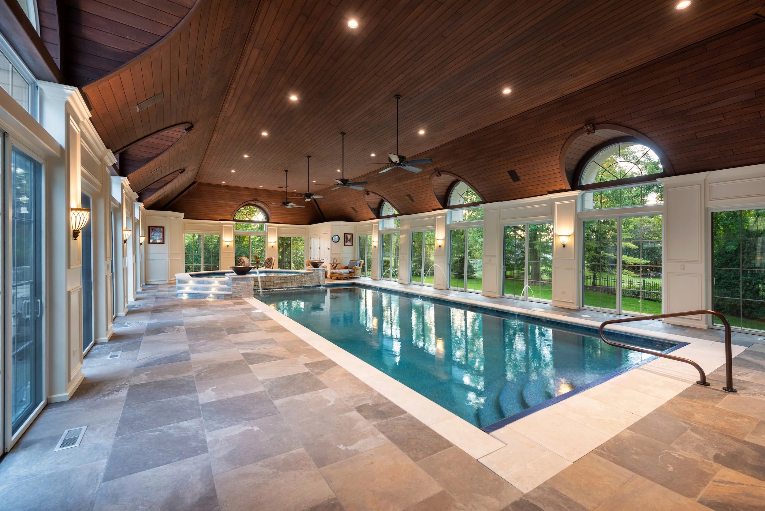Luxurious indoor pool with stone tile flooring, vaulted wooden ceiling, large windows, and elegant lighting.