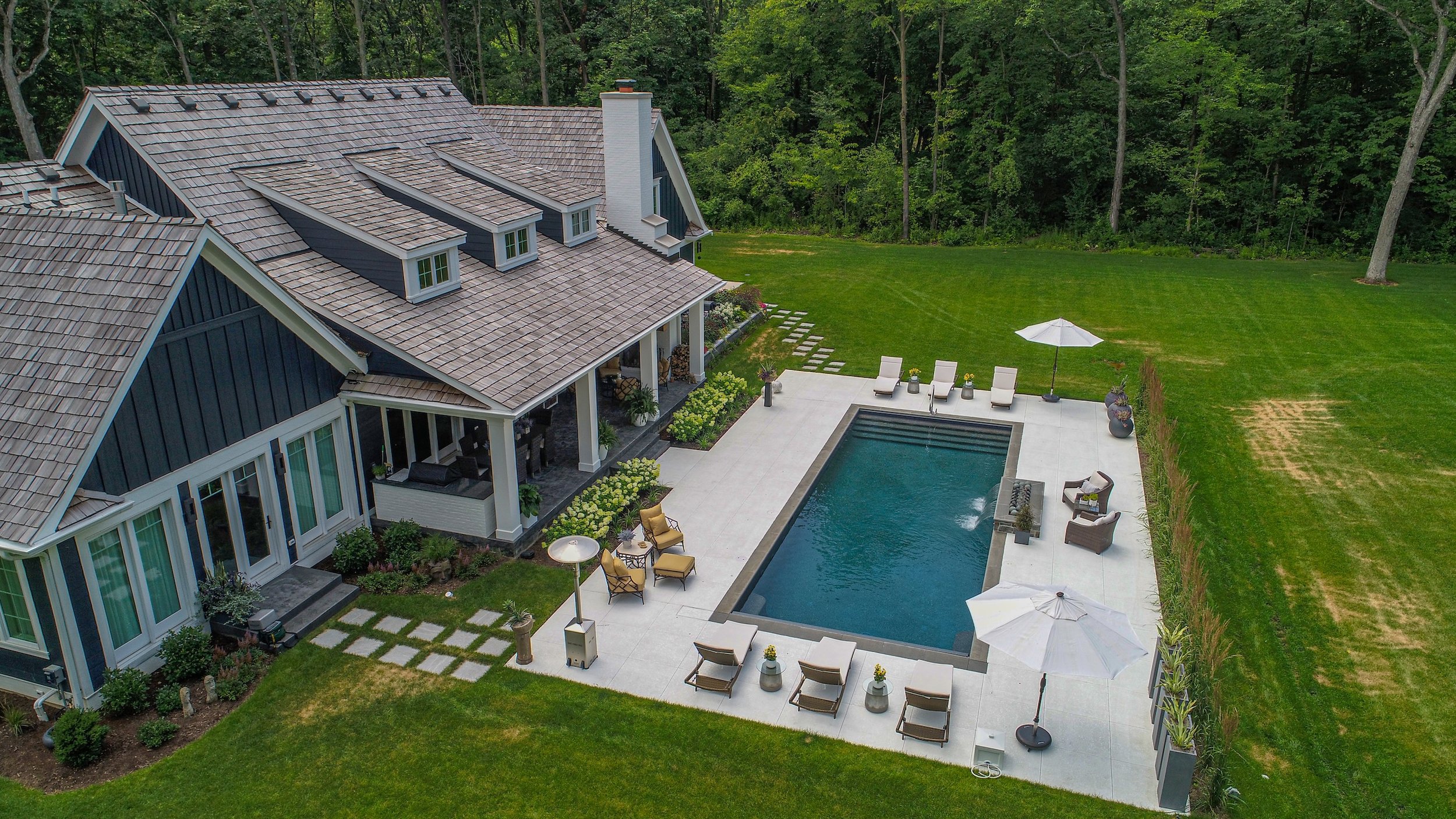 Aerial view of a modern house with a large backyard, featuring a rectangular swimming pool surrounded by a concrete patio. The patio has lounge chairs, tables, and umbrellas. The house has a gabled roof and is surrounded by a well-maintained lawn and