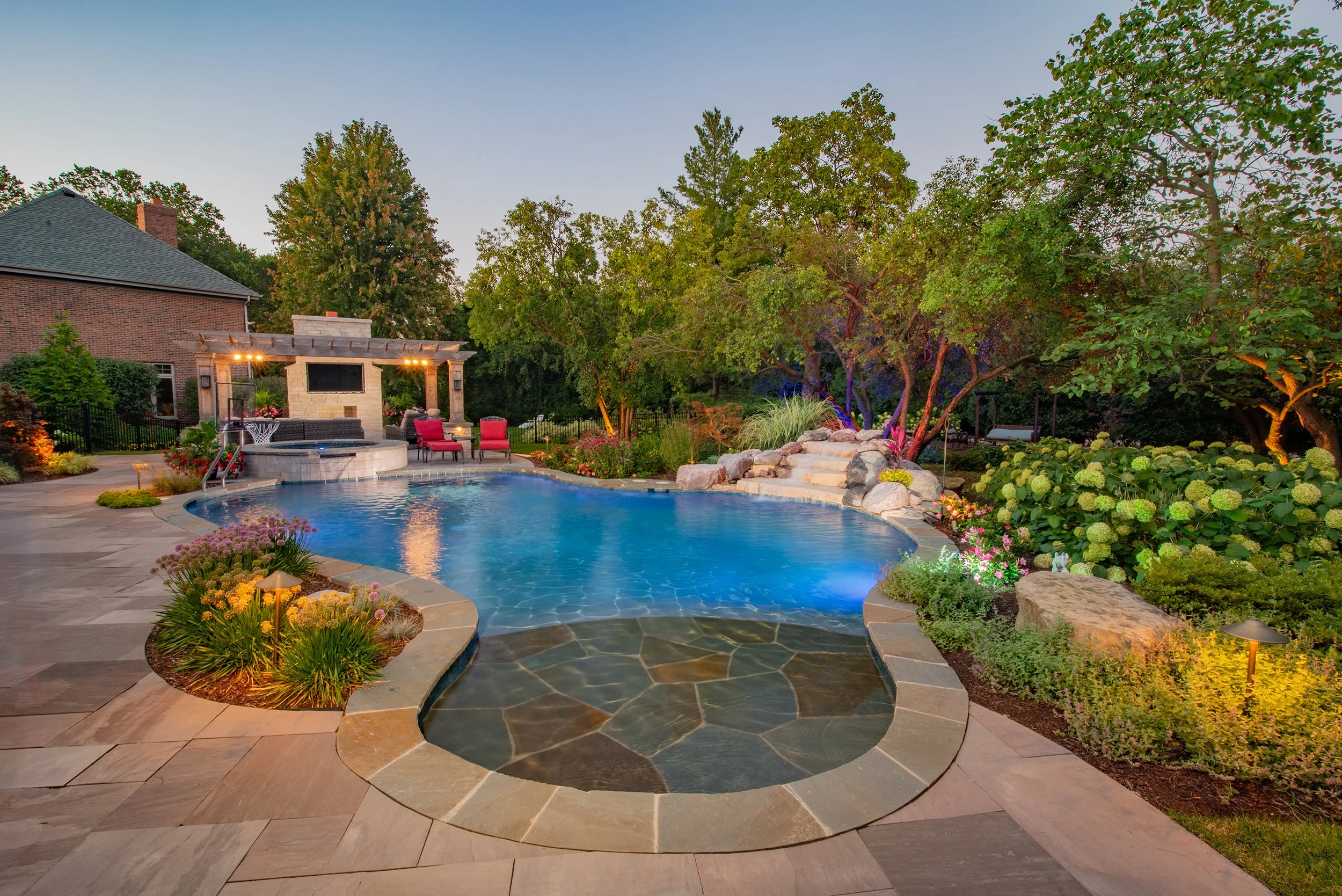 Luxurious backyard with a swimming pool, water features, lush garden, lounge chairs, and a covered patio with a fireplace, surrounded by trees and evening lighting.