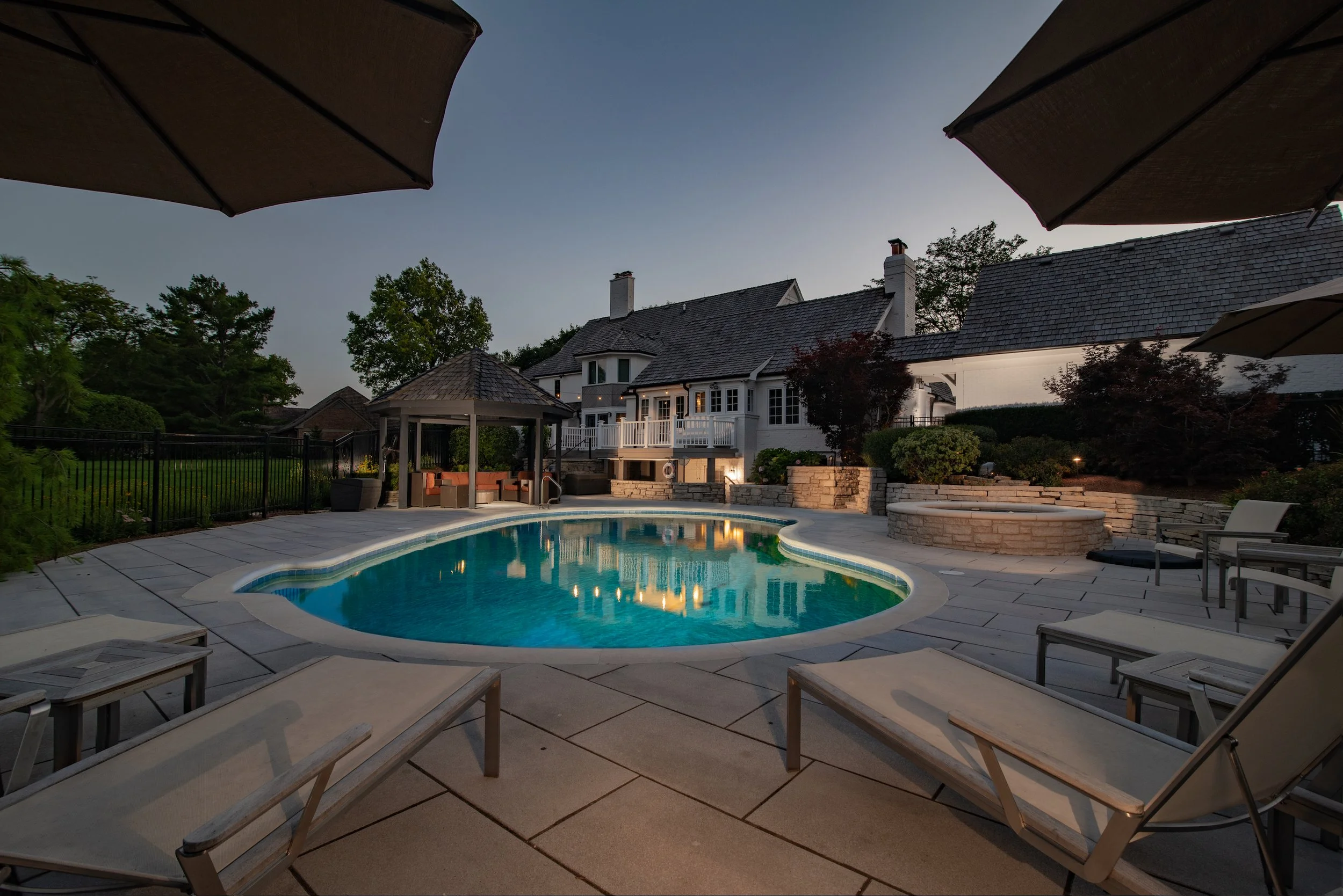 Luxurious backyard with a swimming pool, poolside loungers, a gazebo, and a large house in the background at dusk.