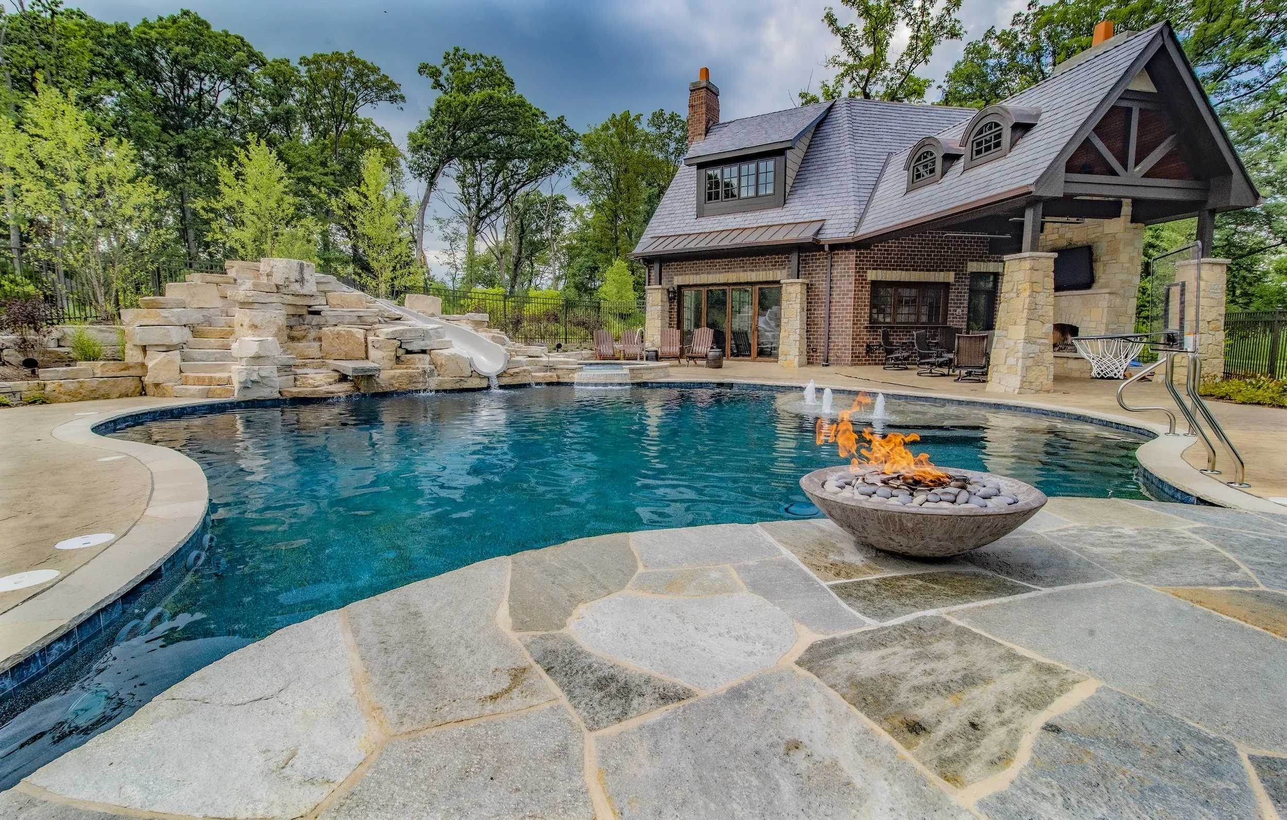Luxury backyard with a swimming pool, stone waterfall, waterslide, fire pit, and brick house surrounded by trees.