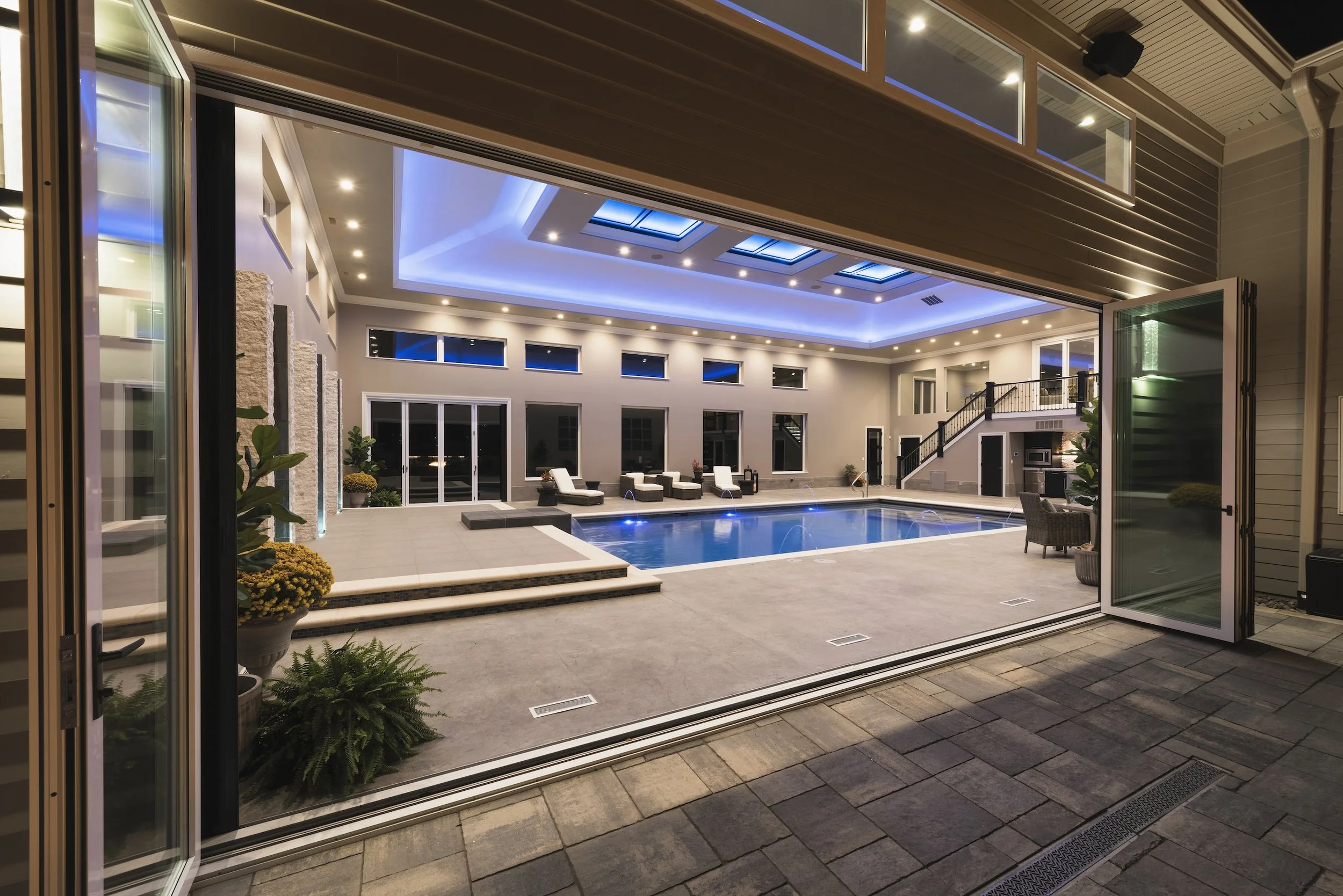 Luxury indoor swimming pool with modern lounge chairs, large windows, stylish lighting, and open sliding glass doors.