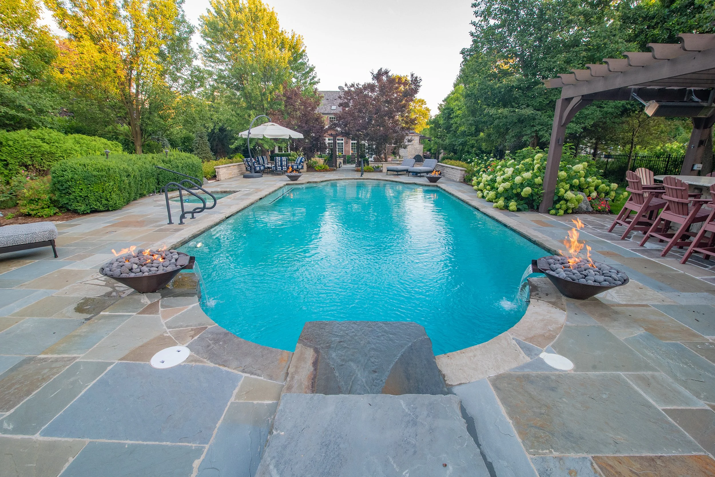 Luxurious backyard swimming pool with fire features, surrounded by greenery, patio furniture, and a pergola with seating.