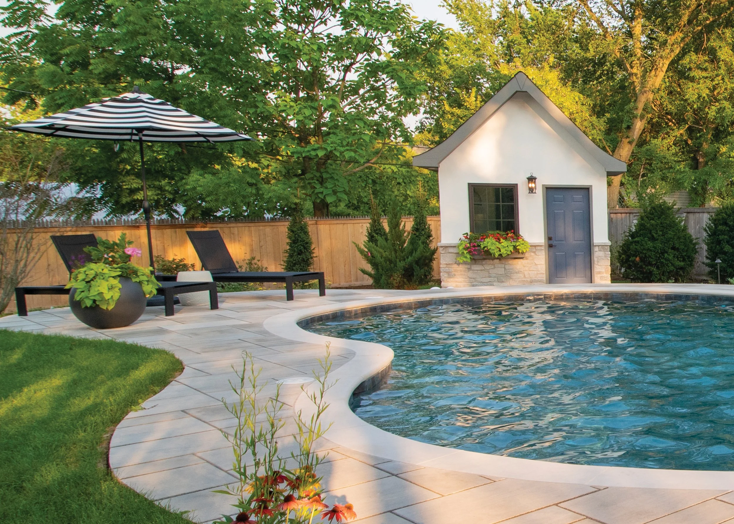 Backyard with a swimming pool, two lounge chairs, black and white striped umbrella, grassy area, small white pool house, potted plants, and trees.