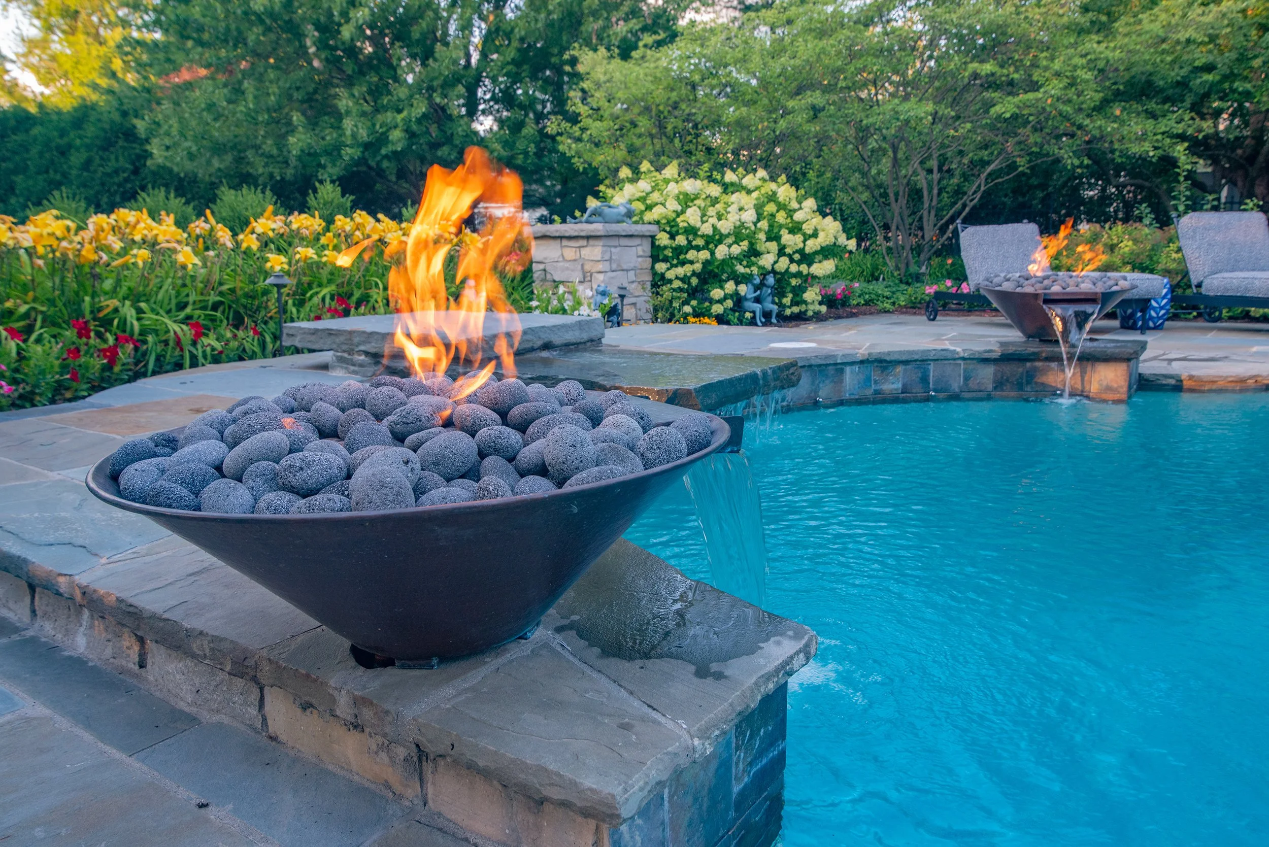 Outdoor pool area with a stone edge, featuring a decorative fire bowl with lava rocks and flames, surrounded by lush garden foliage and seating.