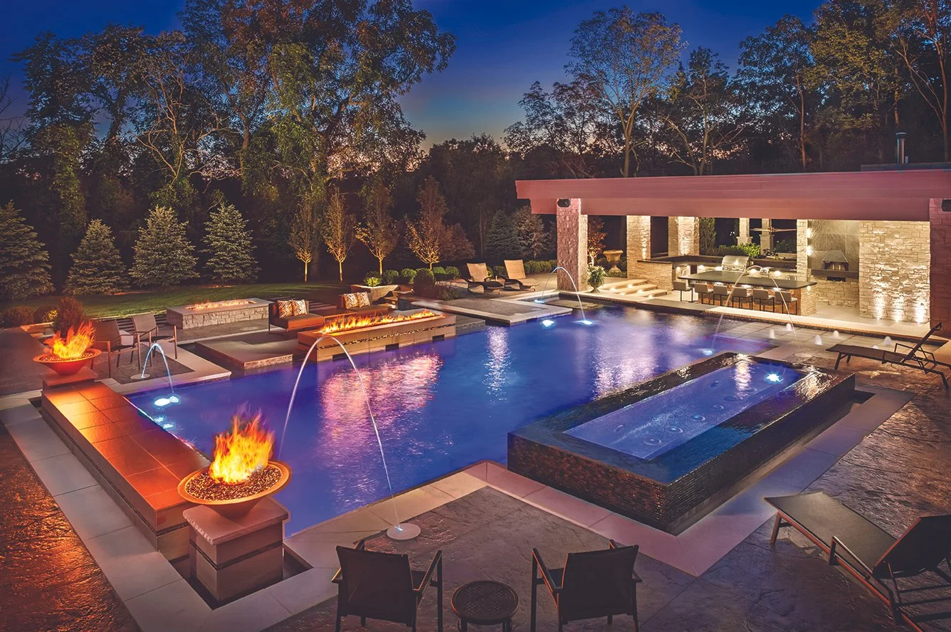 Luxurious backyard with illuminated swimming pool, fire pits, modern seating area, and outdoor kitchen at dusk.