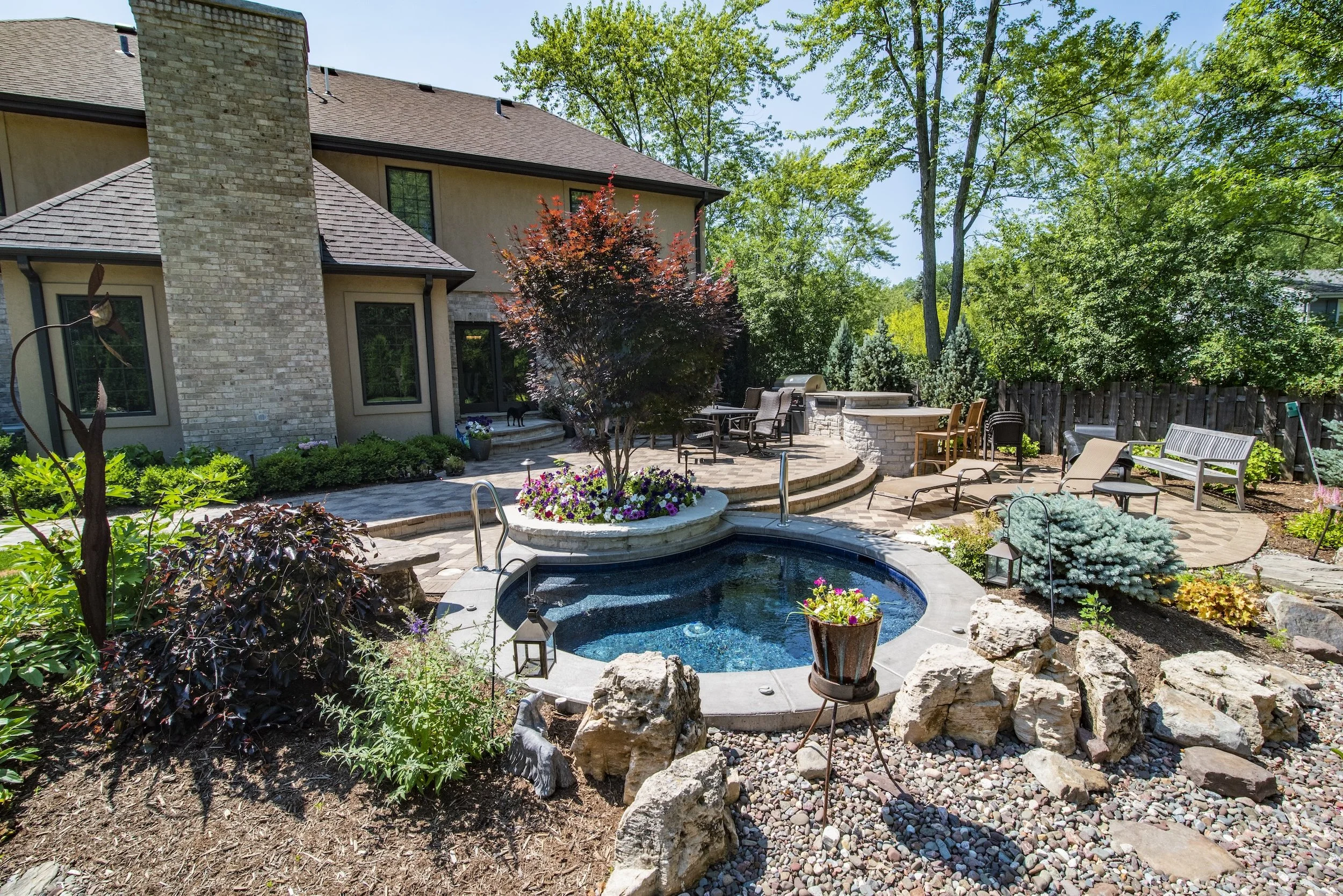 Backyard with a small circular pool, surrounded by rocks and plants, outdoor seating area, lawn chairs, a patio with a stone fireplace, and lush greenery.