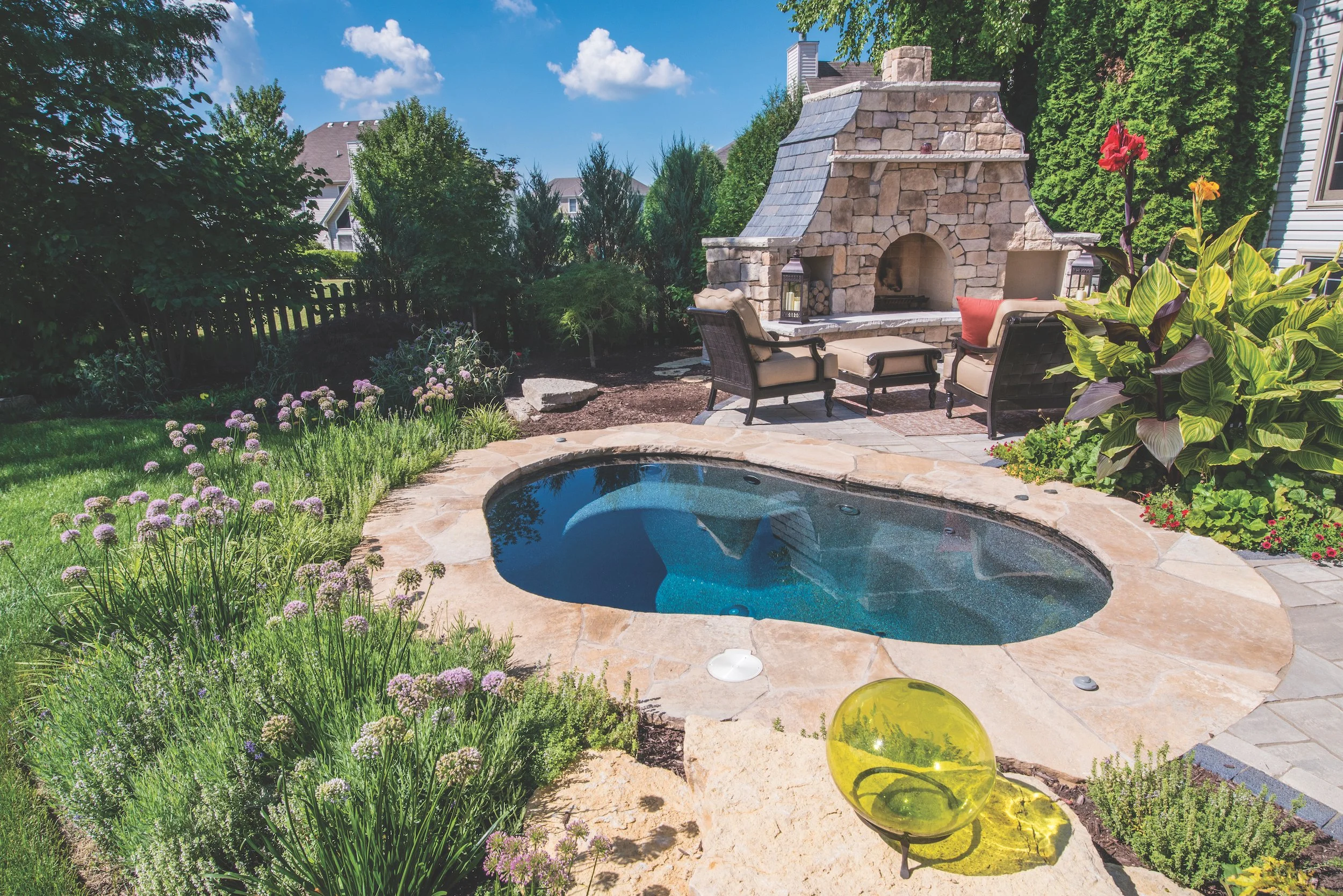 Backyard with a small pool, stone fireplace, patio chairs, and lush landscaping with flowers and plants.