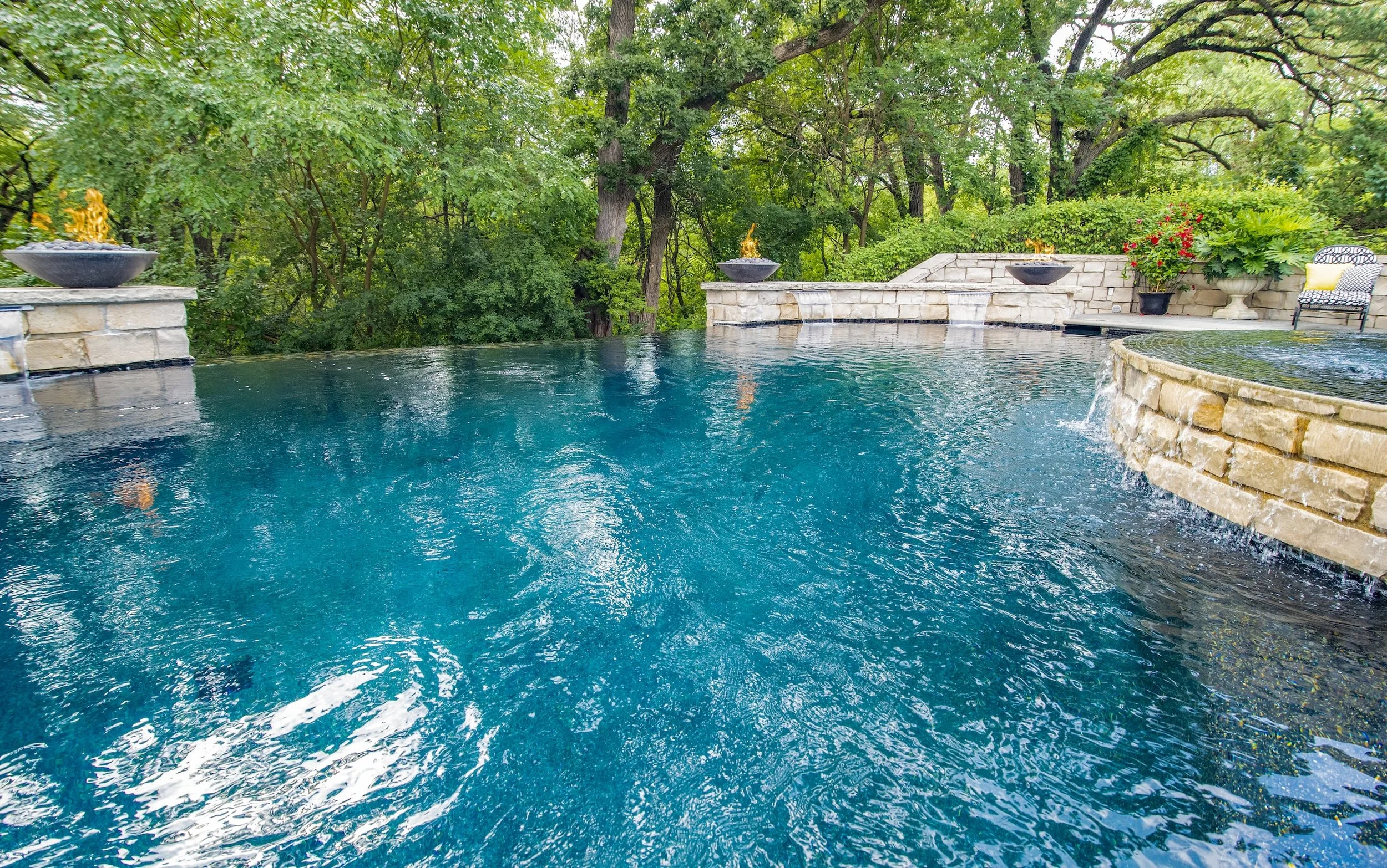 Luxurious infinity pool with stone accents, lush greenery, waterfalls, and fire bowls in a backyard setting.