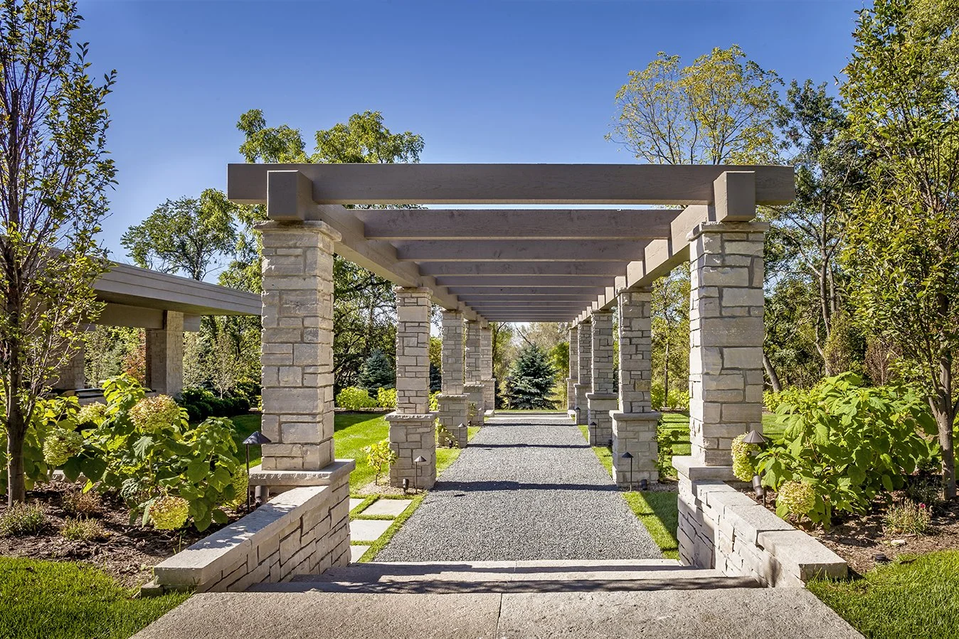 Stone pergola with columns in a garden setting, surrounded by greenery and trees.