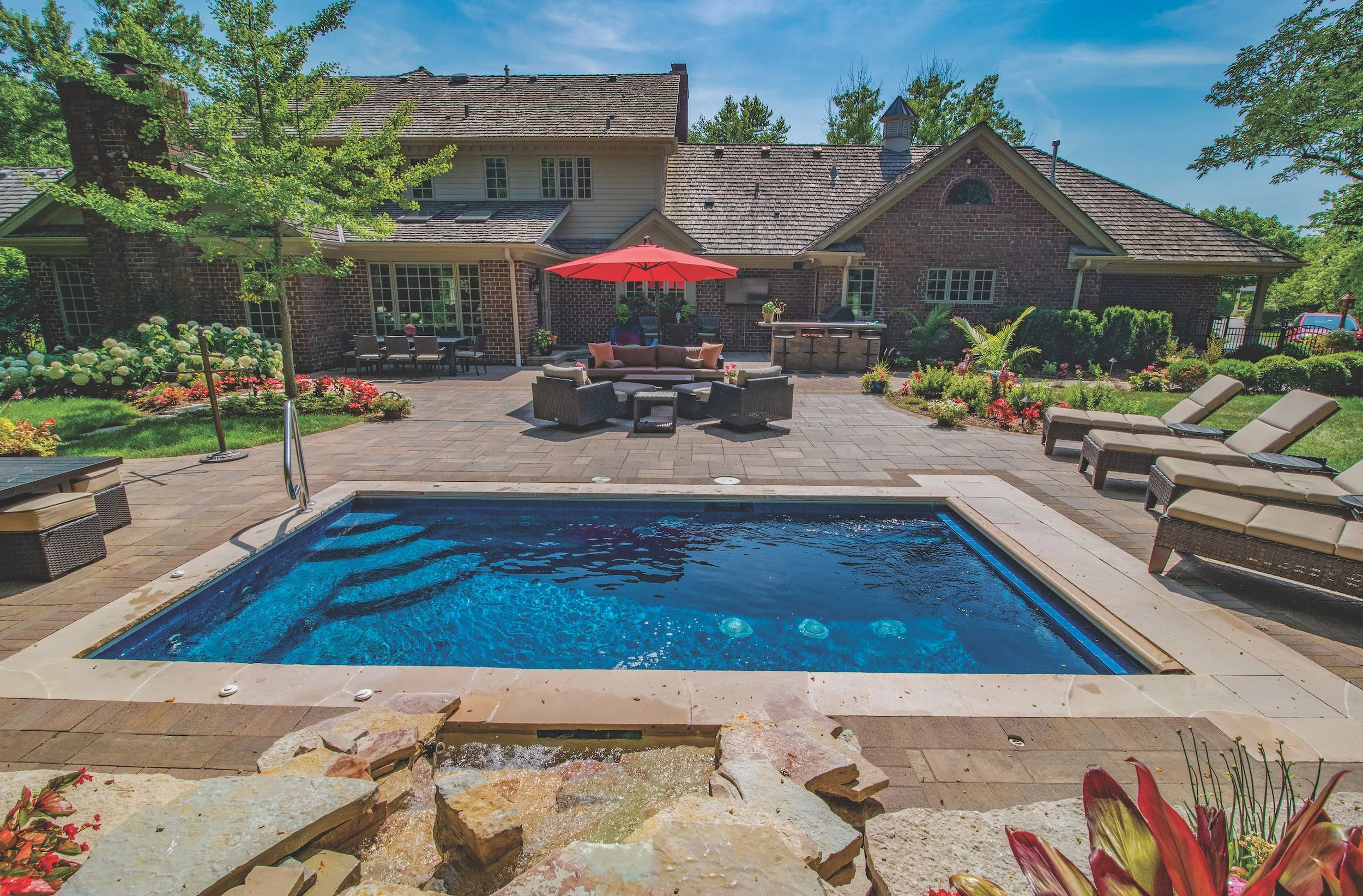 Backyard with a swimming pool, patio furniture, and a red umbrella, surrounded by a well-landscaped garden and a large house in the background.