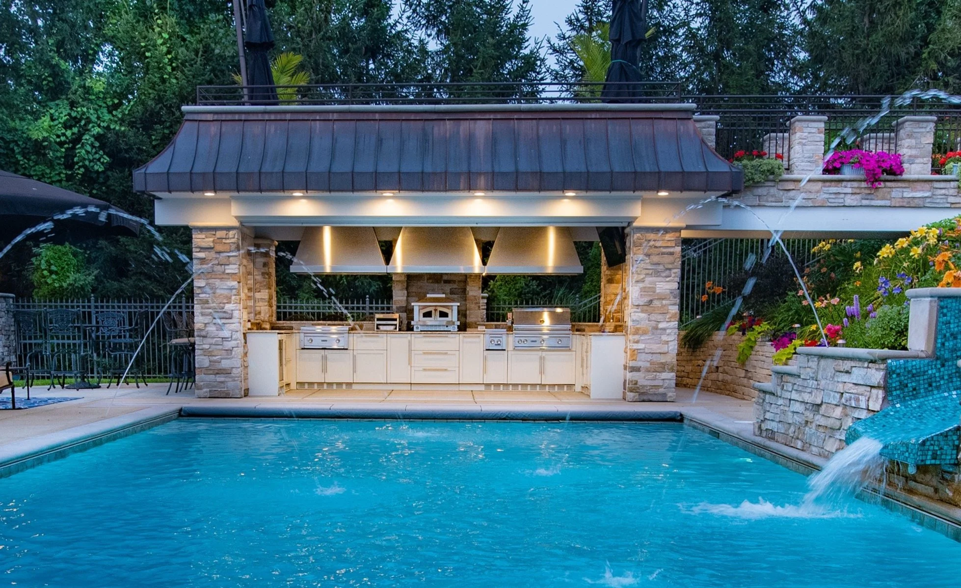 Outdoor kitchen and grill area by a pool with water fountains, surrounded by stone and brick structures and colorful flowers.