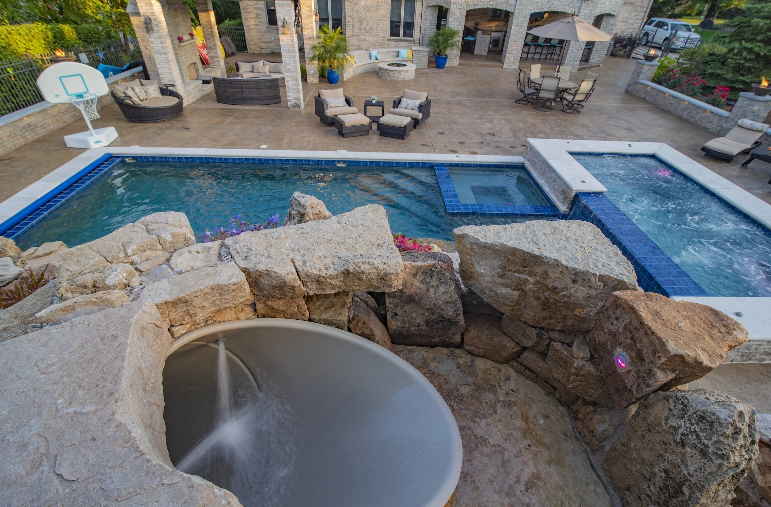Luxury backyard with pool, hot tub, water slide, basketball hoop, stone patio, lounge chairs, covered seating area, fire pit, and outdoor kitchen.