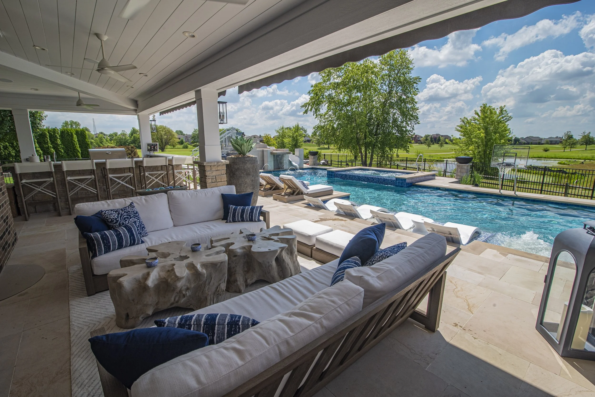 Luxurious outdoor patio with a couch, decorative pillows, wooden table, and a swimming pool. Pool area includes lounge chairs and a basketball hoop. Surrounded by green landscape and a clear blue sky.