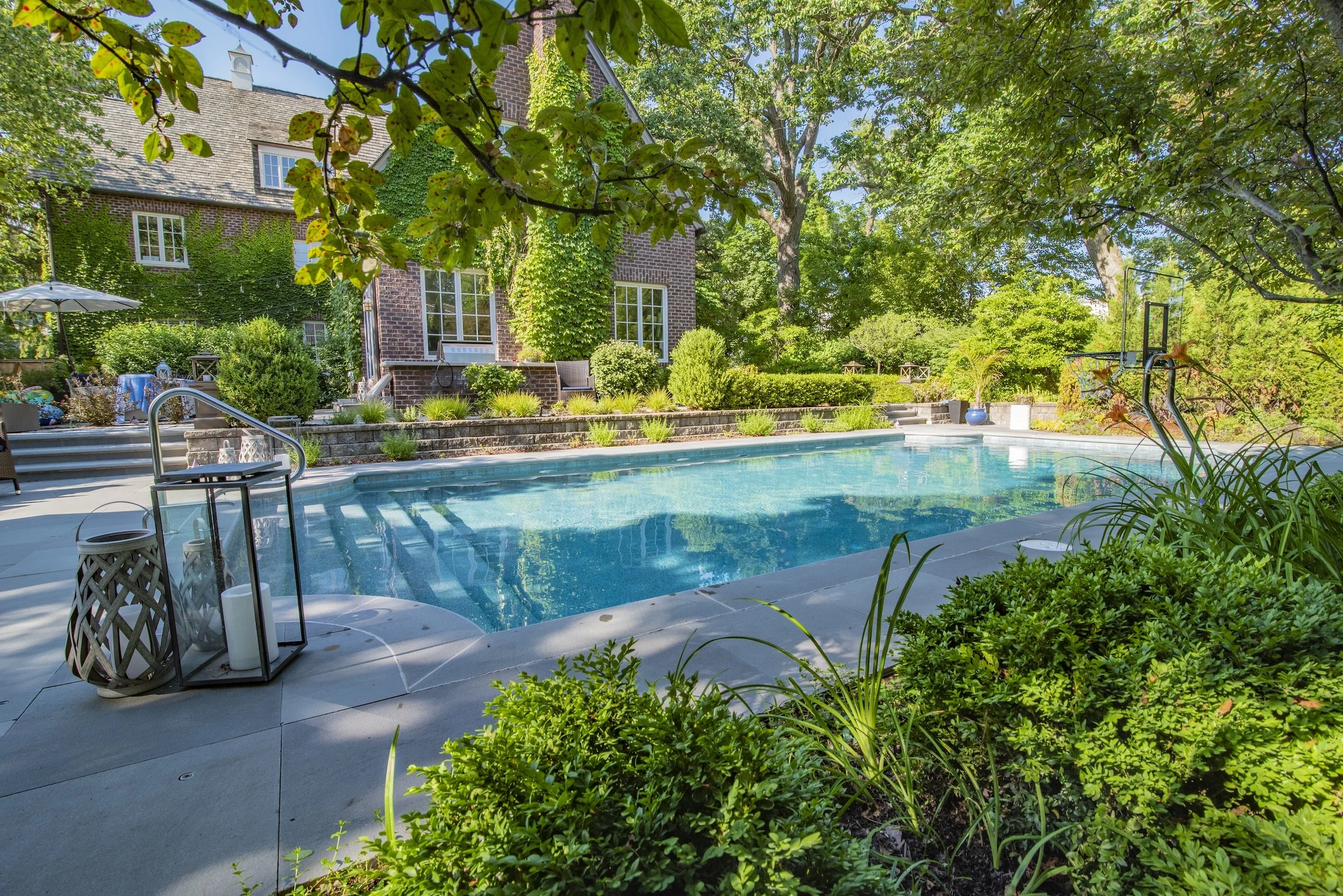 Luxurious backyard with a swimming pool, surrounded by lush greenery and a brick house in the background.