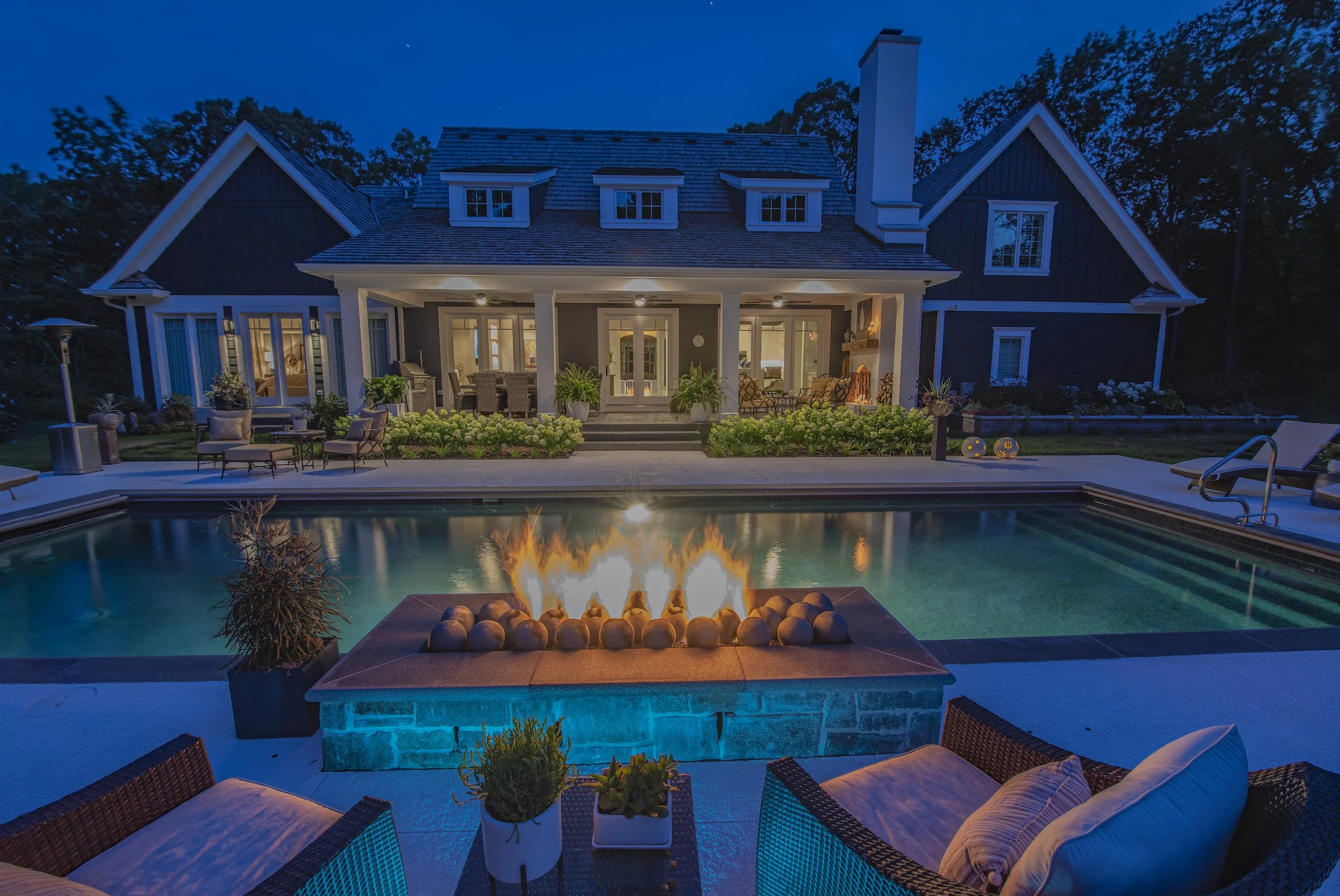 Luxurious backyard with a fire pit, swimming pool, patio seating, and a large house at night.