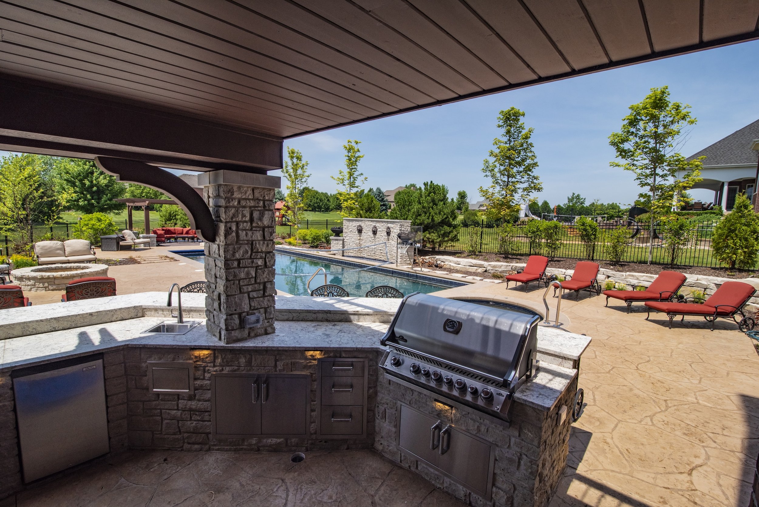 Outdoor kitchen and patio area with a built-in grill, stone countertops, and a view of a swimming pool surrounded by lounge chairs, trees, and a lawn.