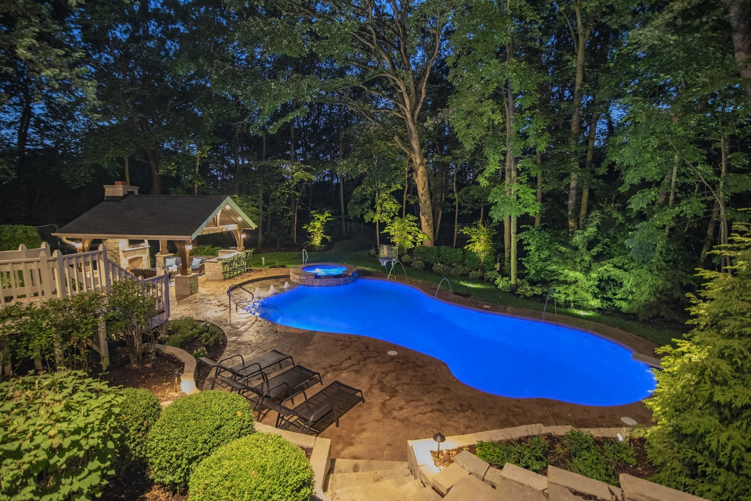Backyard with illuminated swimming pool and hot tub, bordered by trees, poolside chairs, and a covered outdoor seating area at night.