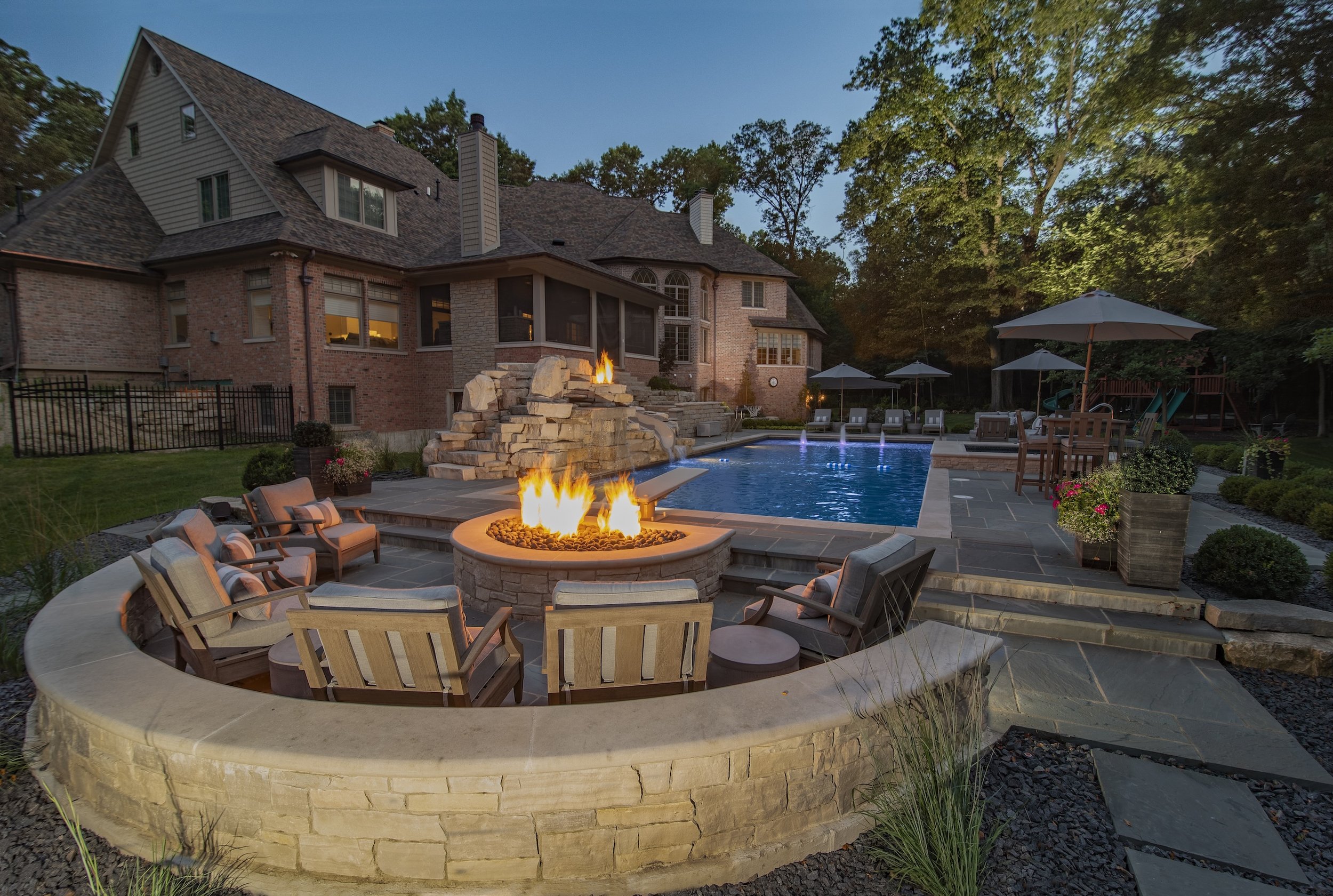 Backyard scene featuring a large house, pool, fire pit, and outdoor seating area surrounded by trees and landscaping at dusk.
